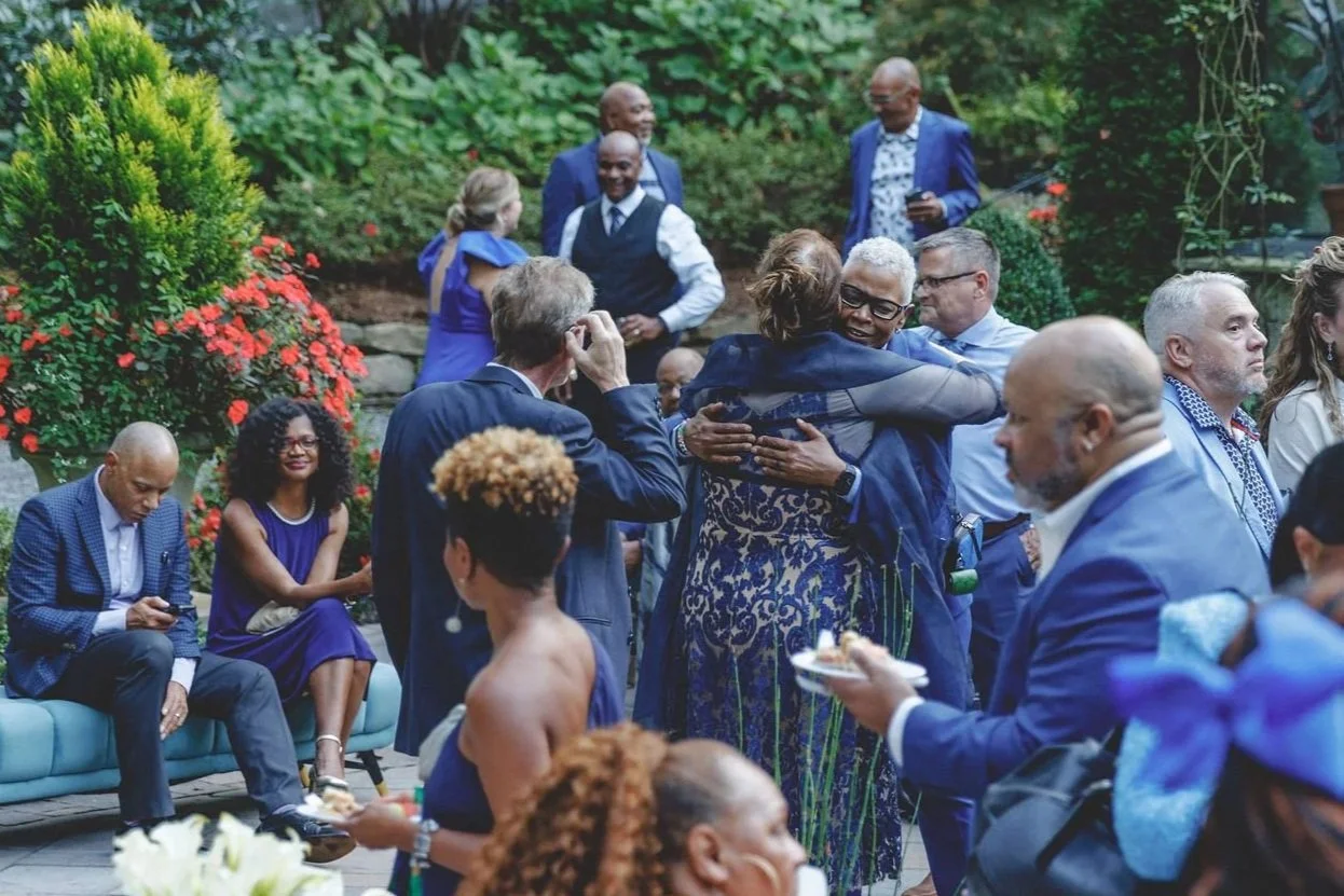 People gathering at an outdoor event, some hugging, some sitting on a couch, others standing and talking, with greenery and flowers in the background.