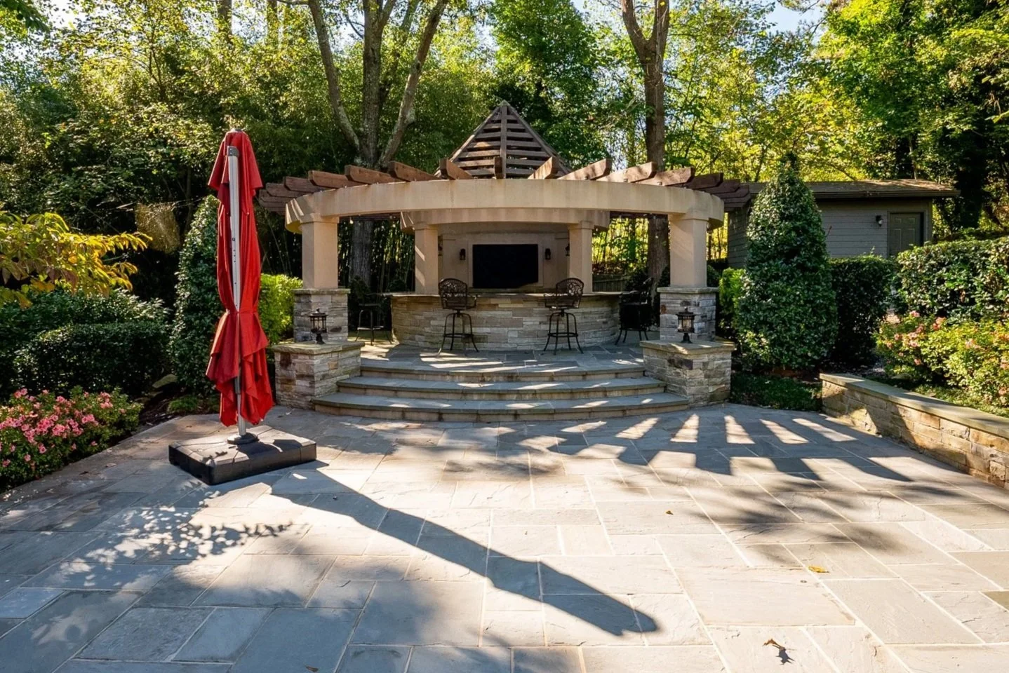 An outdoor patio area with a stone floor, a large closed red umbrella, a built-in outdoor kitchen with bar stools, and lush green trees and bushes in the background.