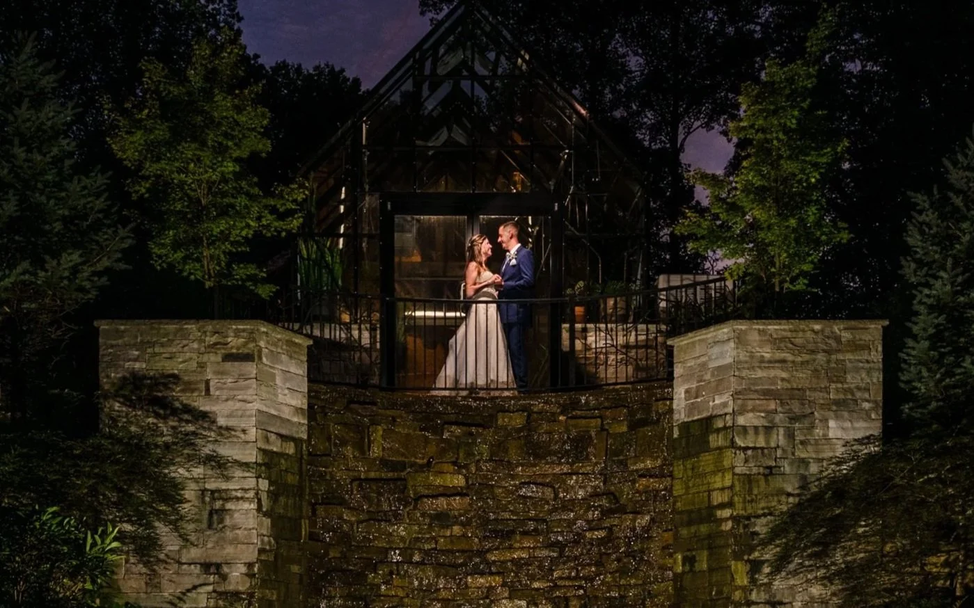 A wedding couple stands on a glass greenhouse balcony at night, holding hands and smiling at each other, surrounded by trees.