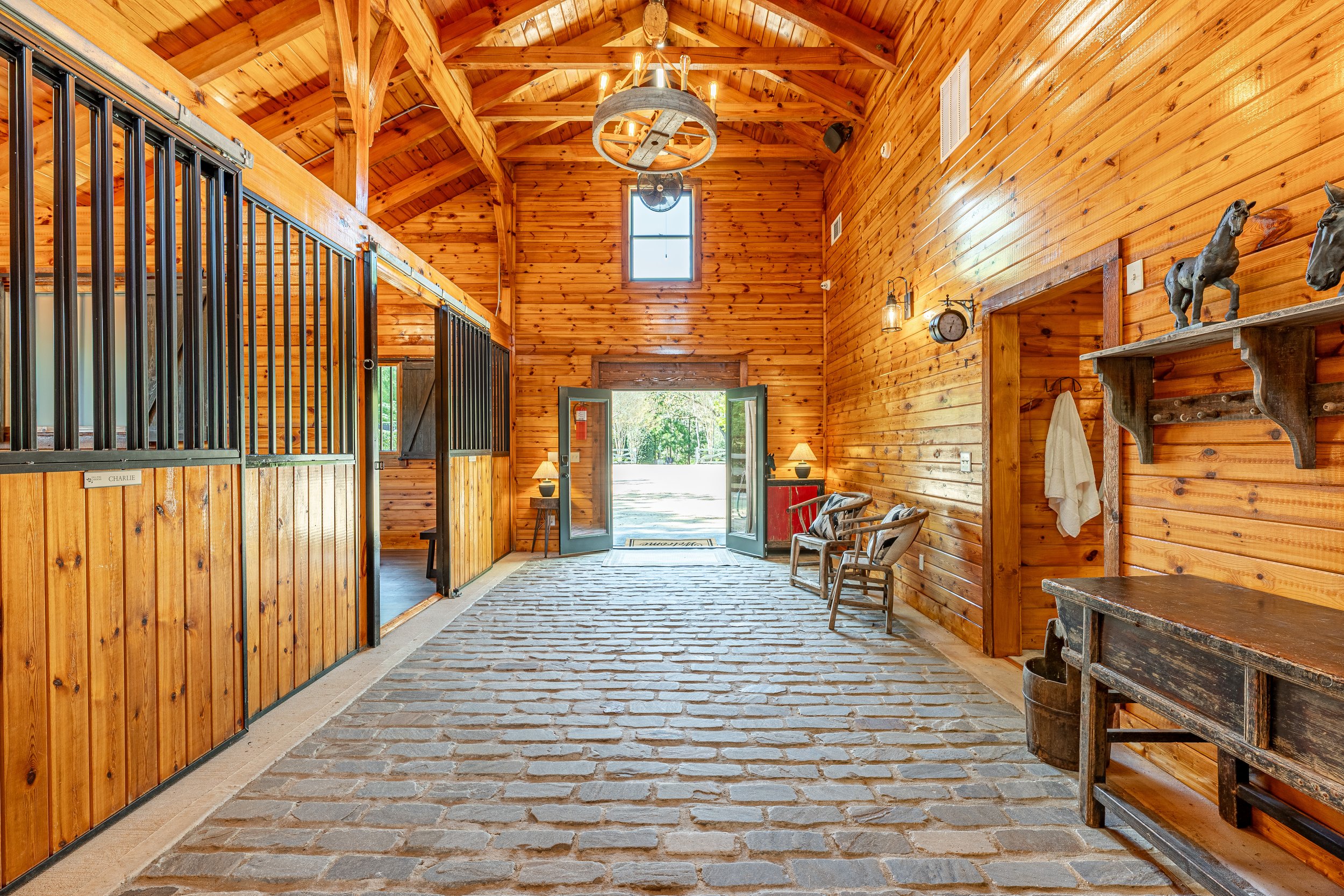 Interior of a horse stable with a rustic wooden design, black metal stall doors on the left, and seating area near open doors at the far end, showing a view of outdoors.