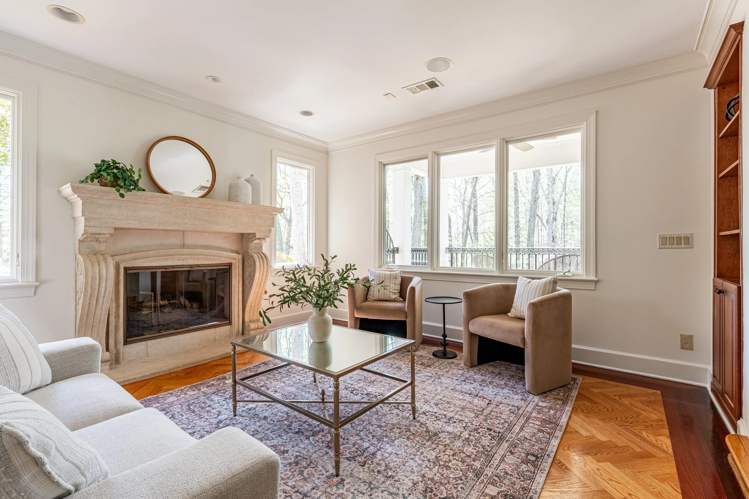 Living room with a fireplace, beige chairs, a glass coffee table, and large windows overlooking a wooded outdoor area.