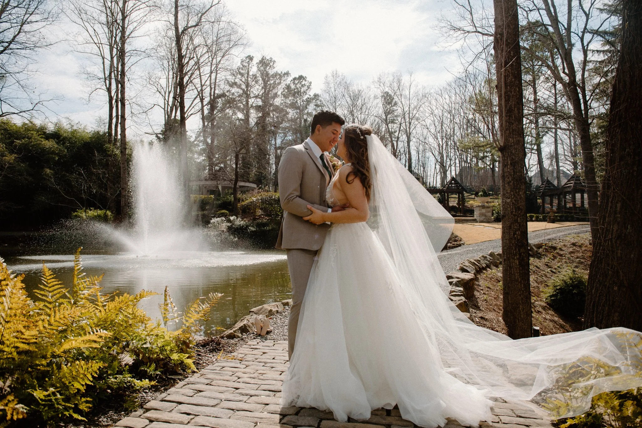 A bride and groom standing together by a pond with a fountain in a park on a sunny day.