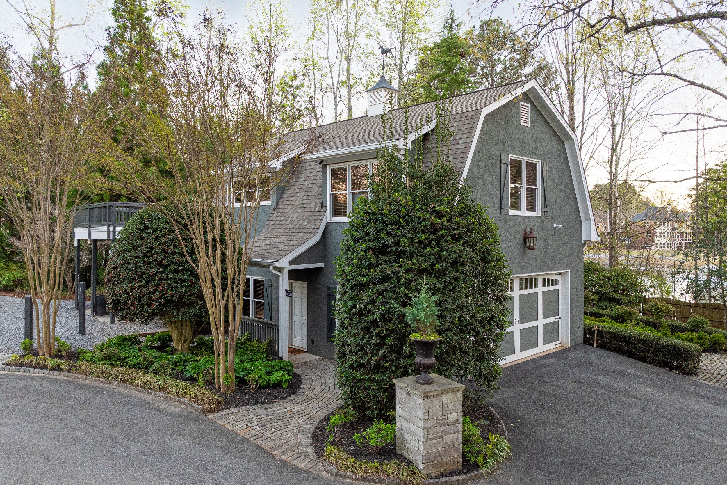 A two-story house with dark gray exterior walls, white trim, and a shingle roof, surrounded by trees and landscaped garden, with a driveway and a garage door.