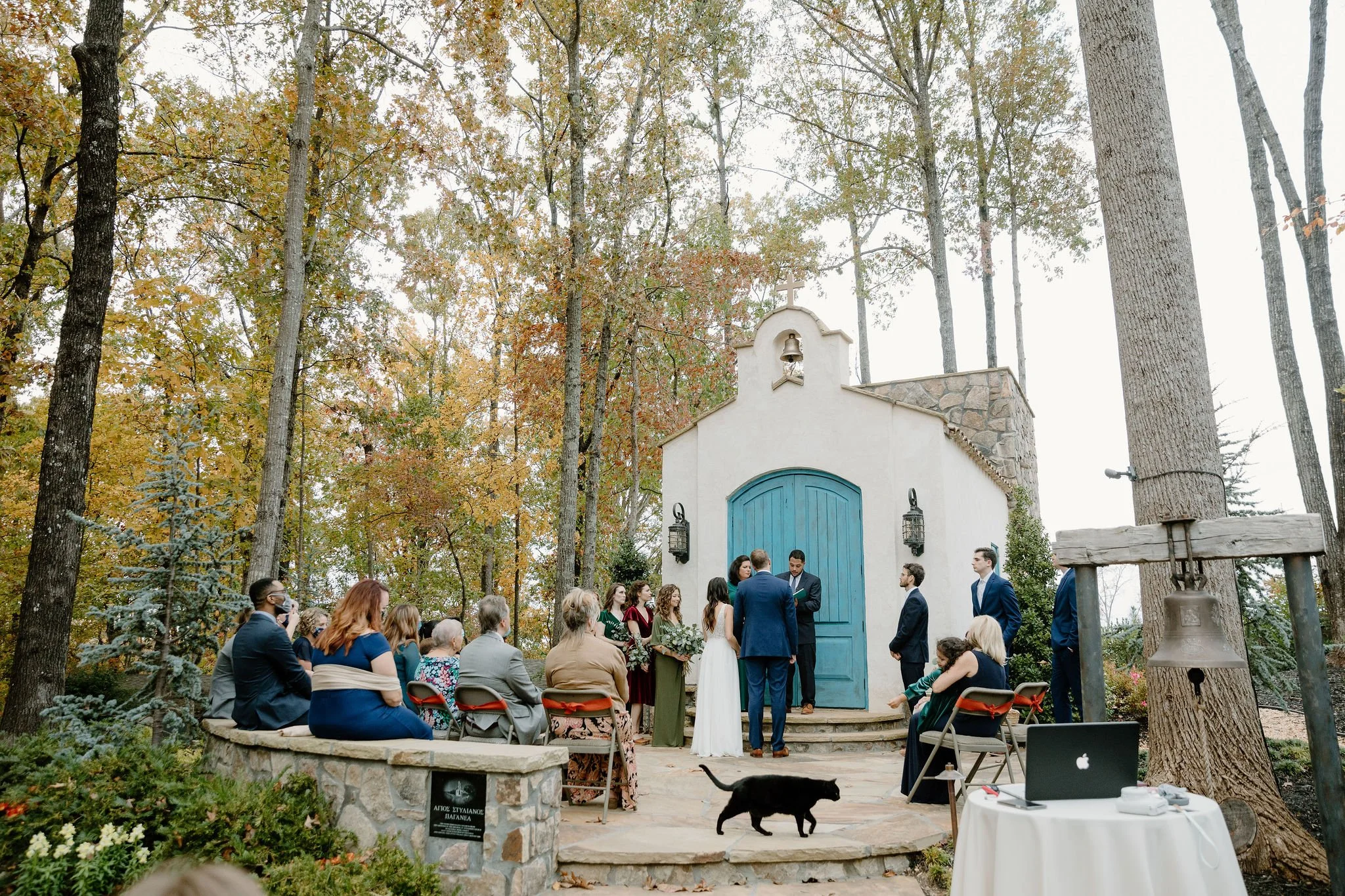 Outdoor wedding ceremony with a small group of people gathered around the officiant and the bride and groom in front of a small white chapel with a blue door and a bell tower, surrounded by tall trees with autumn leaves.