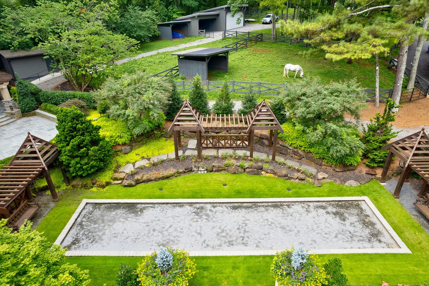 Backyard with a sand volleyball court, green grass, trees, bushes, a fenced horse pasture with a white horse, and a grey shed.