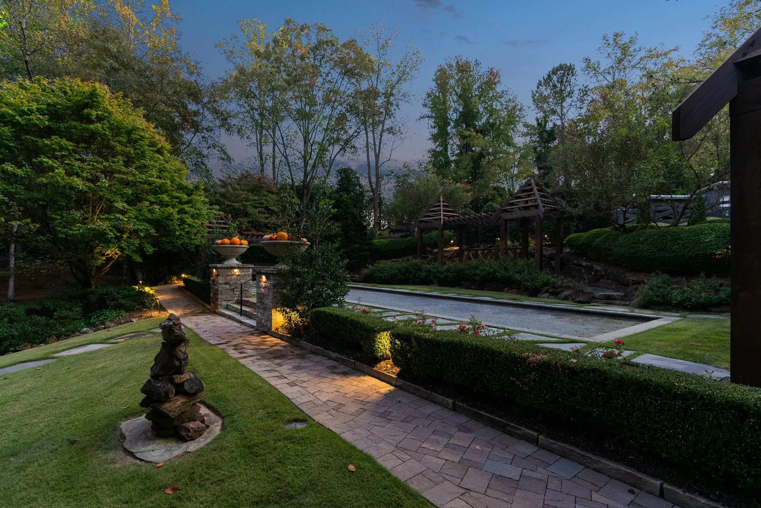 A well-lit backyard garden during dusk features a stone and brick pathway, artistic rock sculpture, neatly trimmed bushes, a water feature, and wooden gazebos among tall trees and flowering plants.