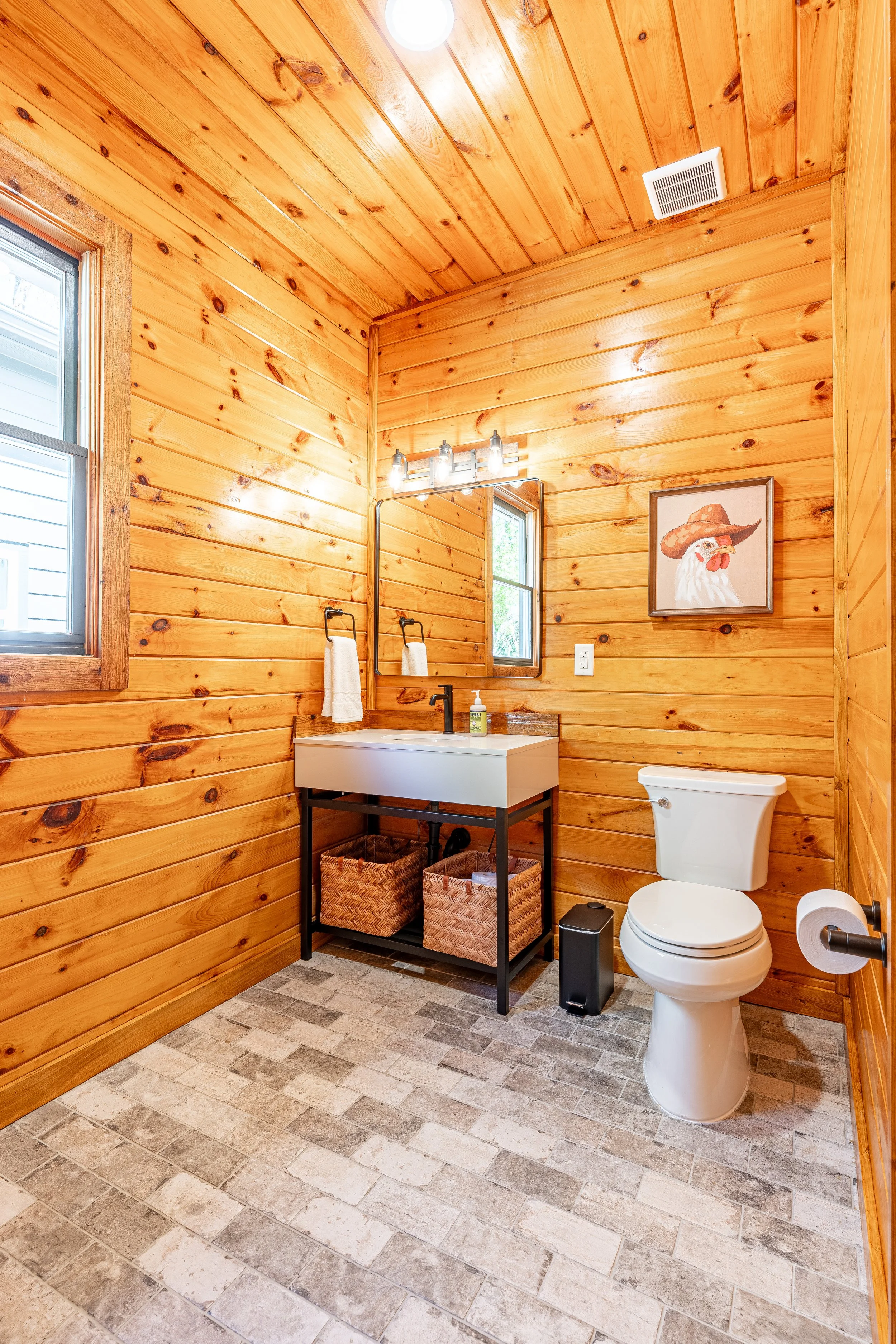 A rustic bathroom with wooden paneled walls and ceiling, a white toilet, a black metal vanity with two woven baskets underneath, a rectangular mirror, a small window, and a framed picture of a chicken wearing a cowboy hat.