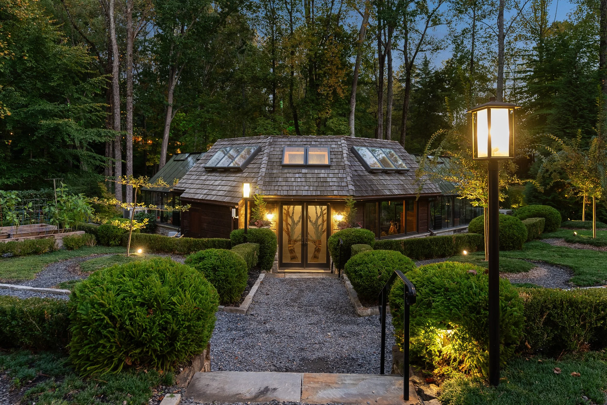 A house with a unique roof surrounded by well-maintained landscaping and outdoor lighting, set in a wooded area at dusk.