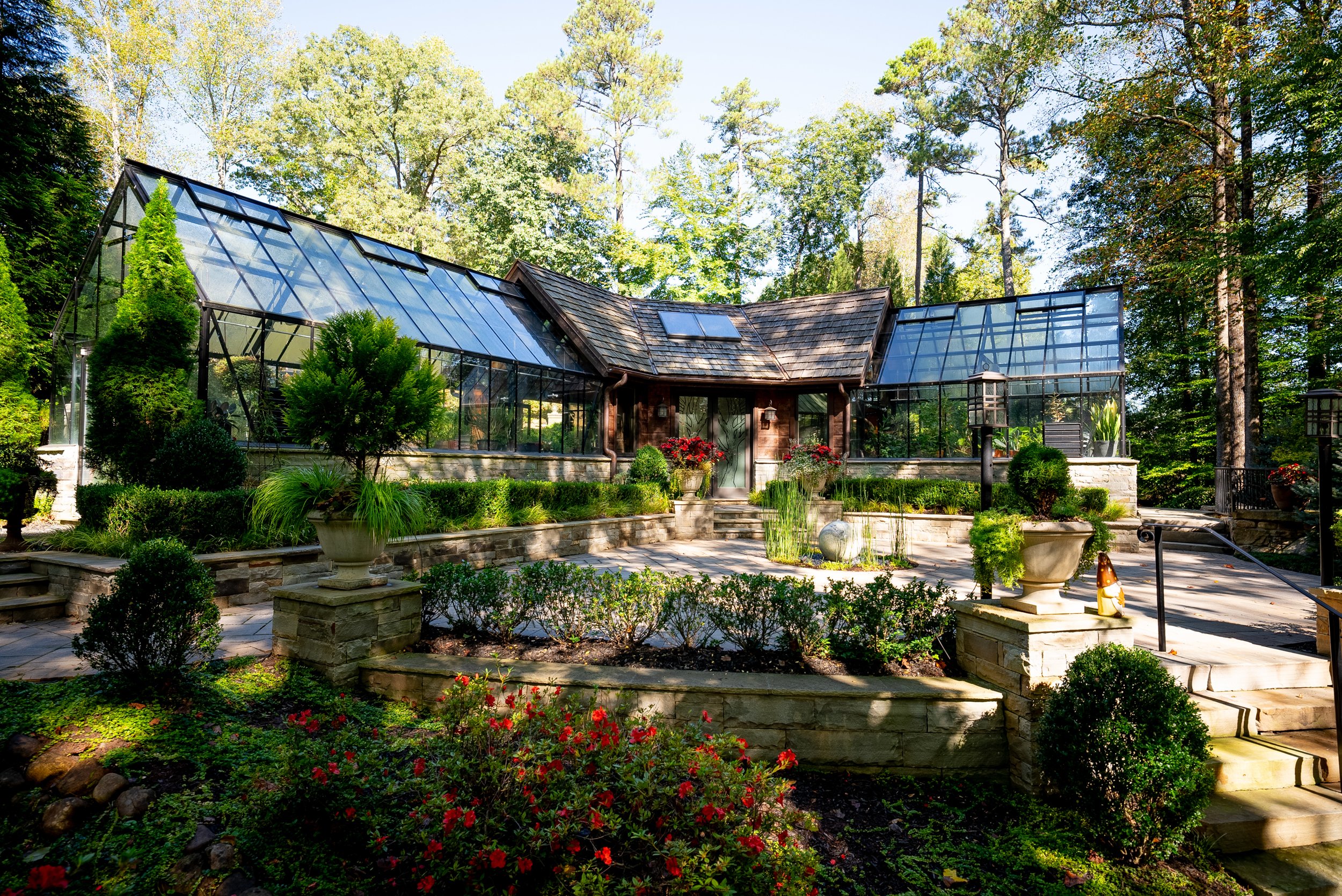 A large glass greenhouse on stone foundation in a lush garden with stairs, trimmed bushes, flowers, and trees.