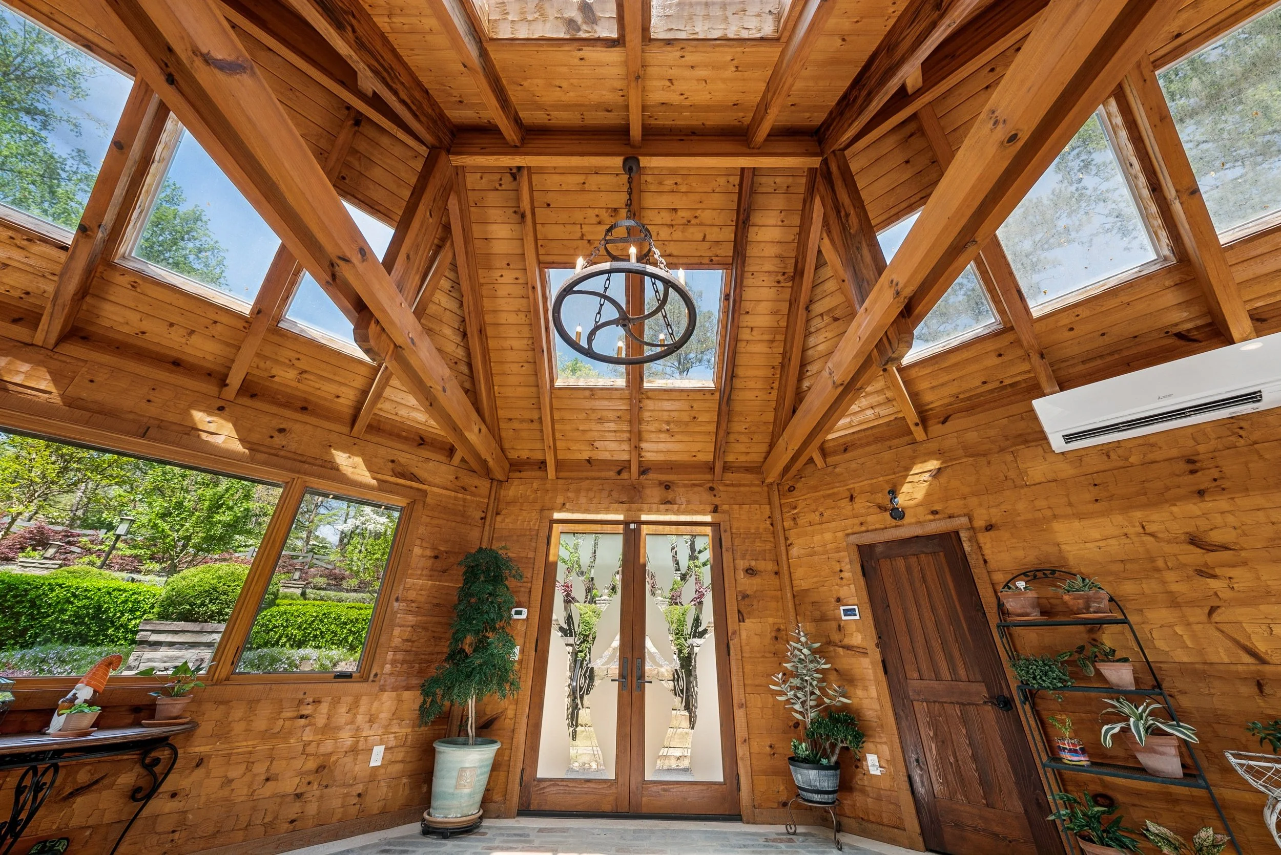 Interior view of a wooden house with high ceiling, multiple skylights, and large windows. The walls and ceiling are made of natural wood, with decorative plants and a chandelier hanging from the ceiling. Double glass doors lead outside to a garden wi