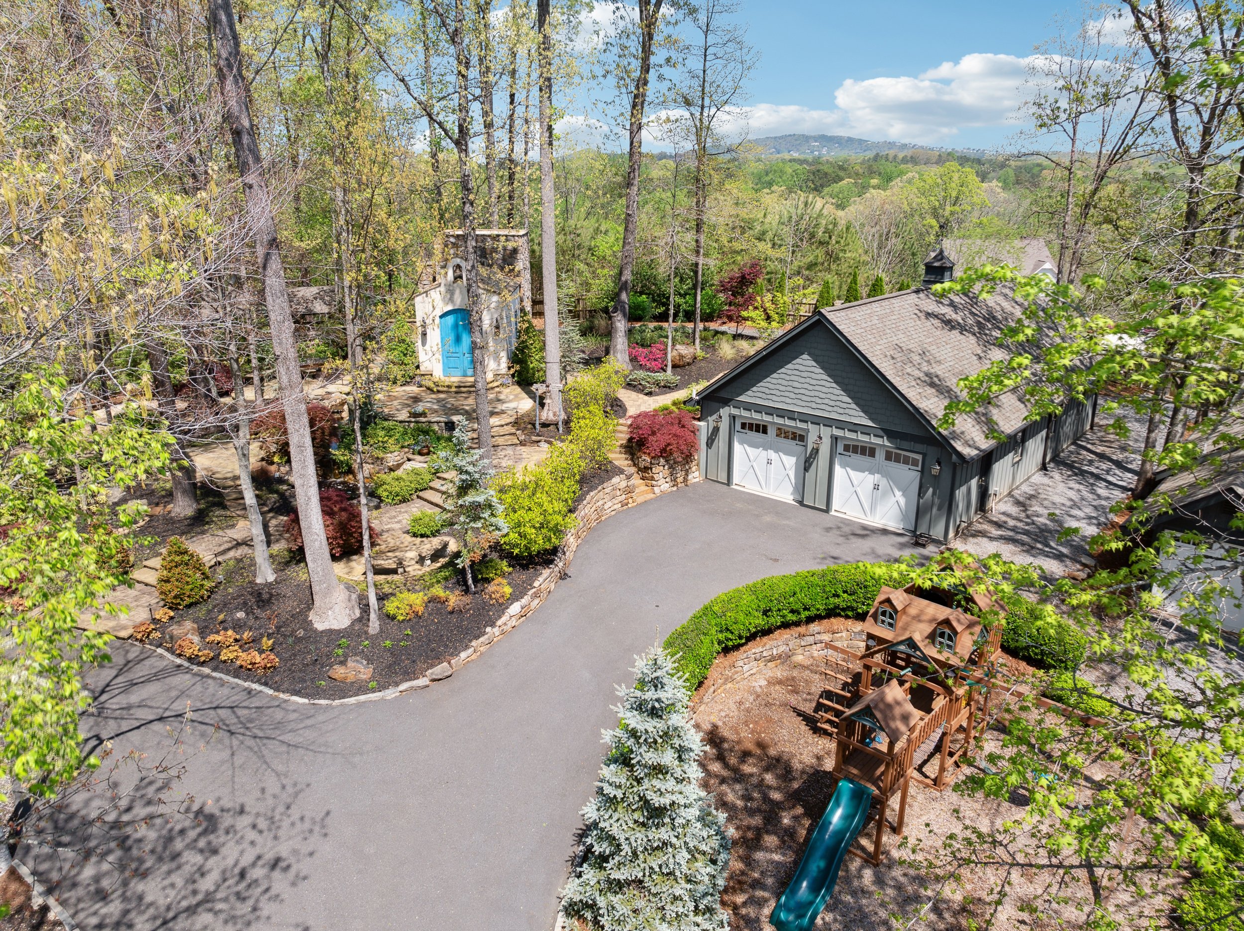 Aerial view of a backyard with a gray garage, a wooden playset with a slide, a landscaped garden with trees and bushes, and a small blue door on a garden shed or structure, surrounded by a driveway and greenery.