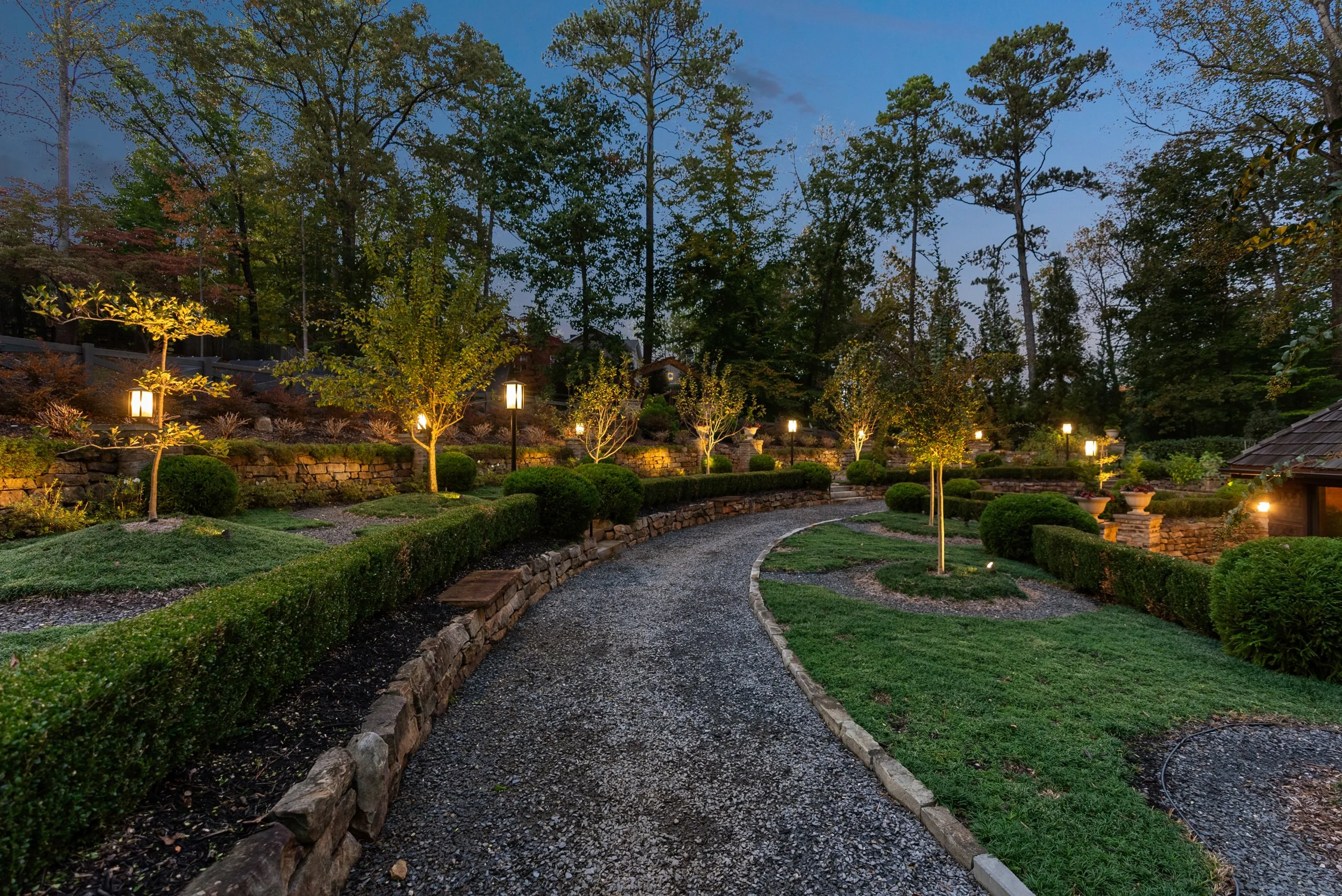 A garden pathway at dusk with stone-edged gravel, surrounded by trimmed bushes, small trees, and decorative lighting along the path, with larger trees and a hill in the background.