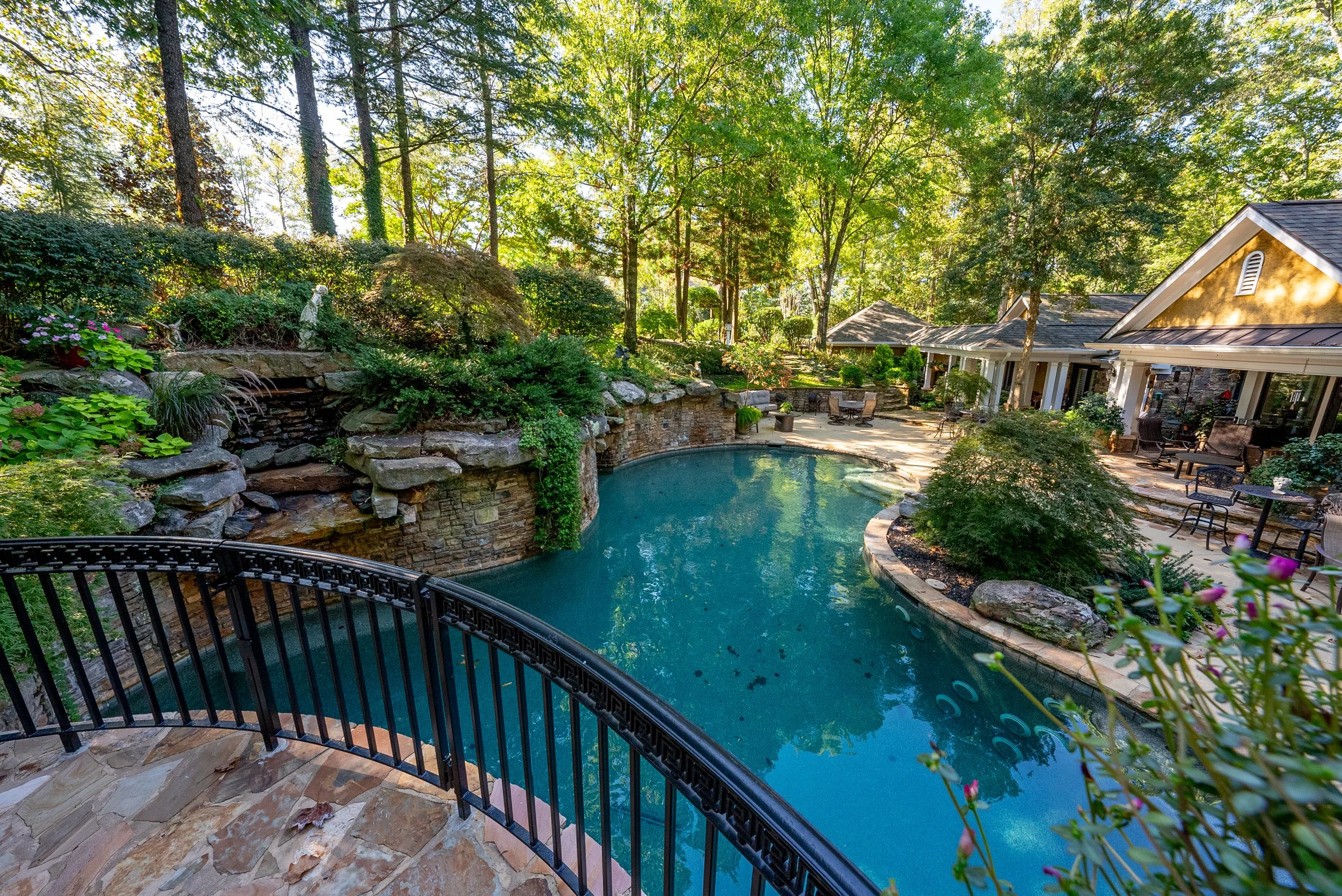 A backyard with a swimming pool surrounded by trees, a stone patio with outdoor furniture, and a house with a covered porch.