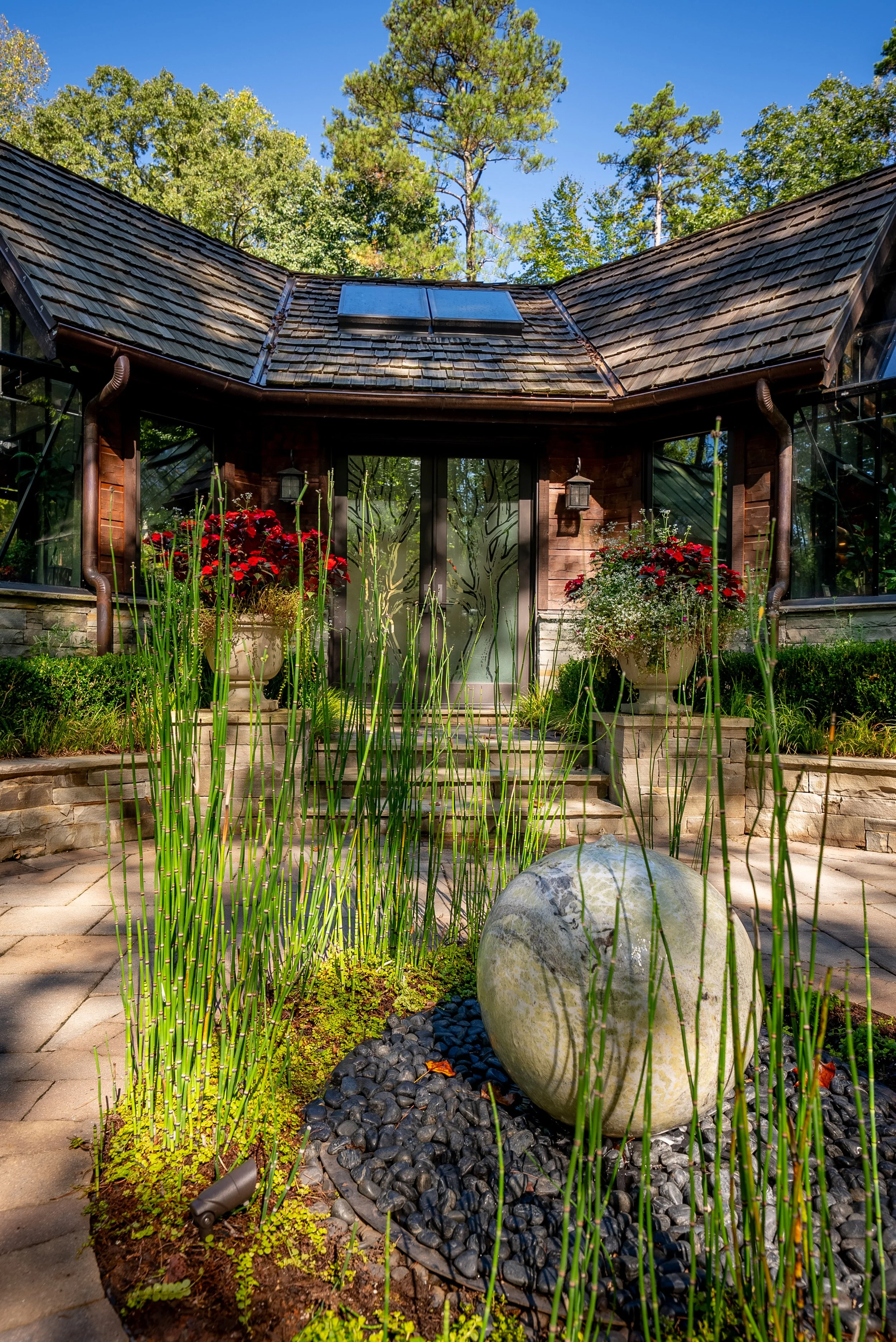 Front view of a house with a stone pathway, large planter pots with red flowers, and a garden with tall grasses in the foreground, trees with green foliage and a blue sky in the background.