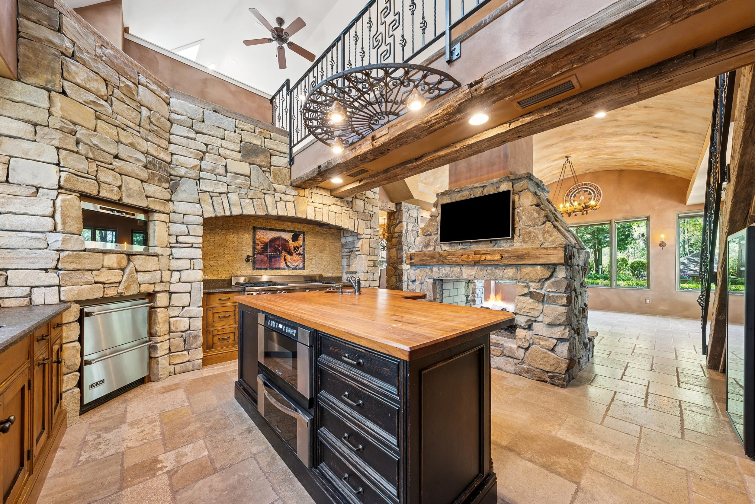 Interior of a rustic kitchen featuring stone walls, a large wooden kitchen island, and a stone fireplace with a mounted television above. There is an upper loft with a decorative wrought iron railing and ceiling fan. Large windows reveal greenery out