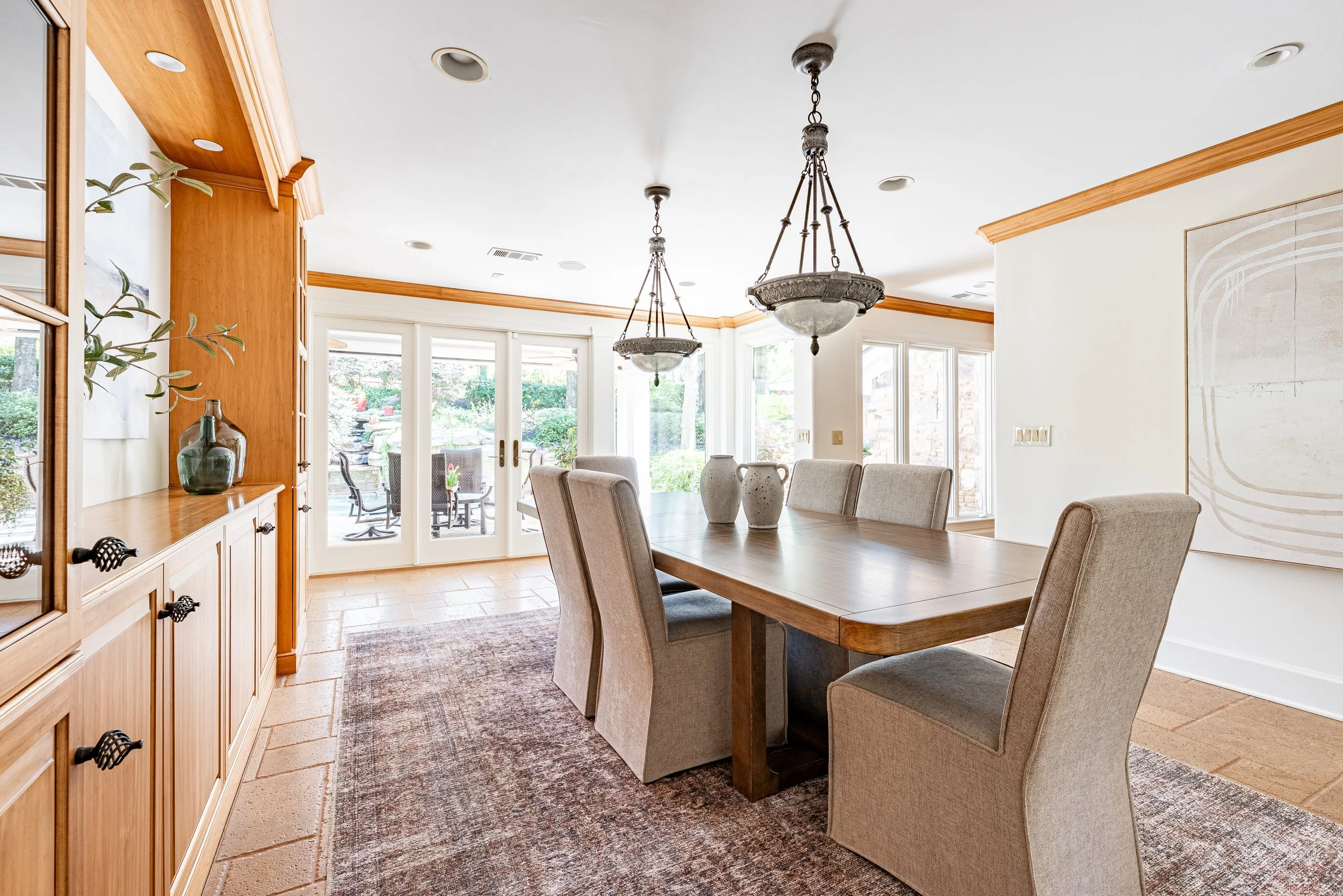 A dining room with a wooden table surrounded by six beige upholstered chairs, a wooden cabinet with decorative items on the left, large glass doors leading to an outdoor patio, and two hanging chandeliers.