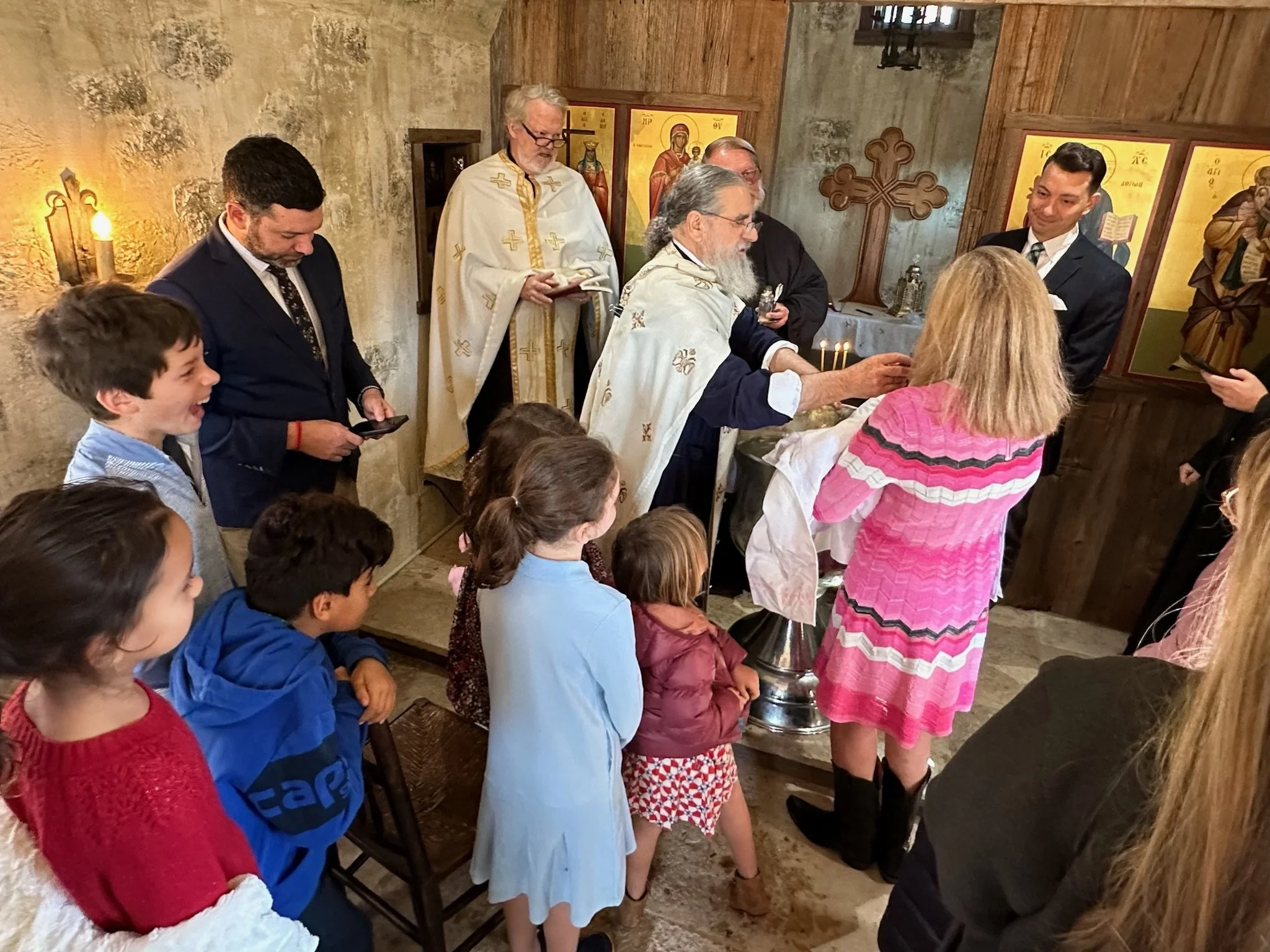 A religious ceremony in an Orthodox church with priests and attendees, including children and adults, near icons and a cross on the wall.