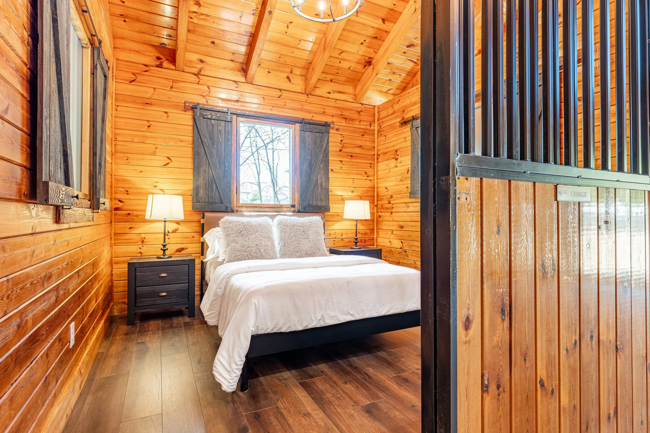 A cozy bedroom with wood-paneled walls and ceiling, a bed with white bedding and fluffy pillows, two side tables with lamps, and a window with dark shutters.