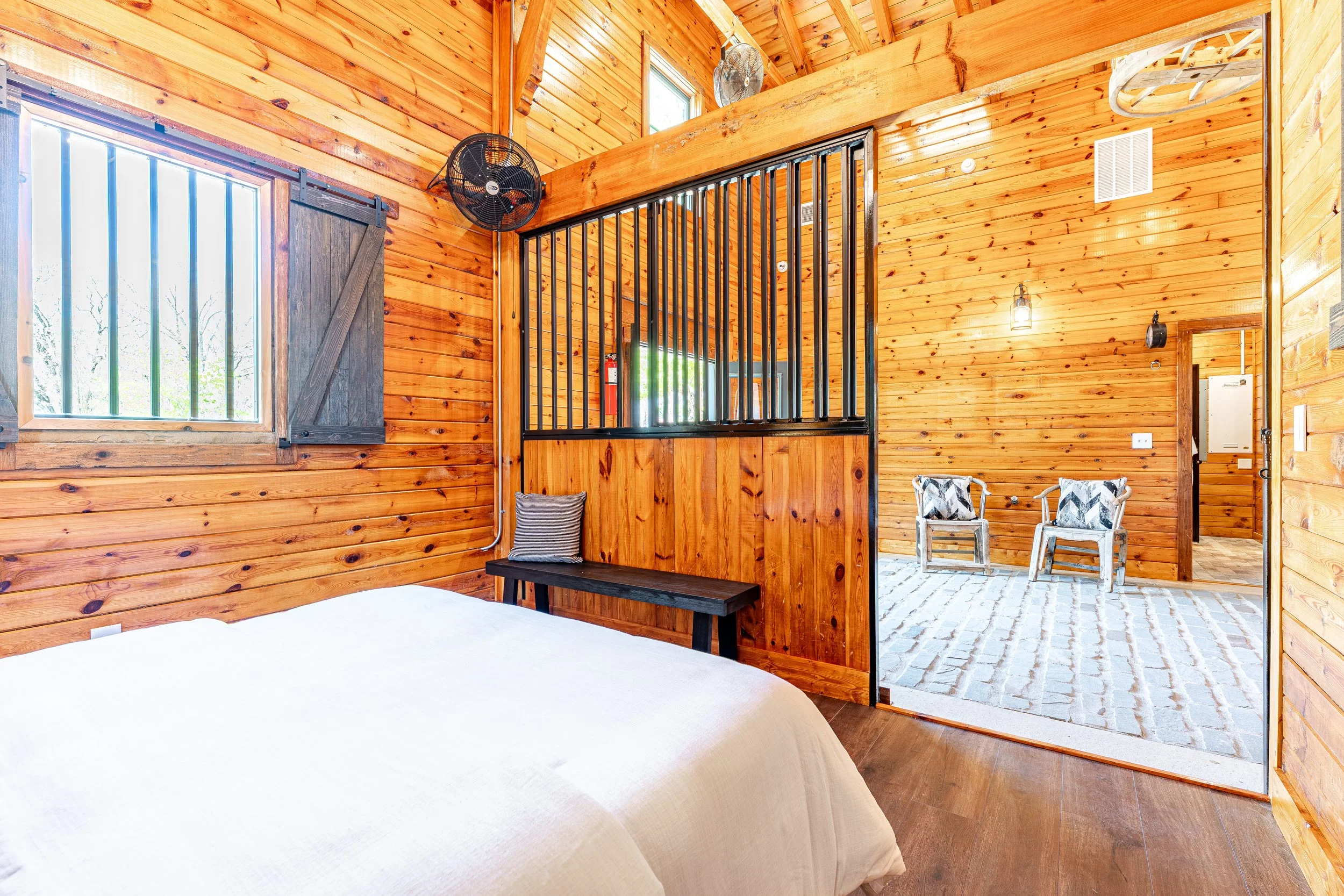 Interior of a wooden cabin bedroom with a white bed, a window with black shutters, a bench with a pillow, and a separated living area with two chairs with patterned cushions, and a door leading outside.