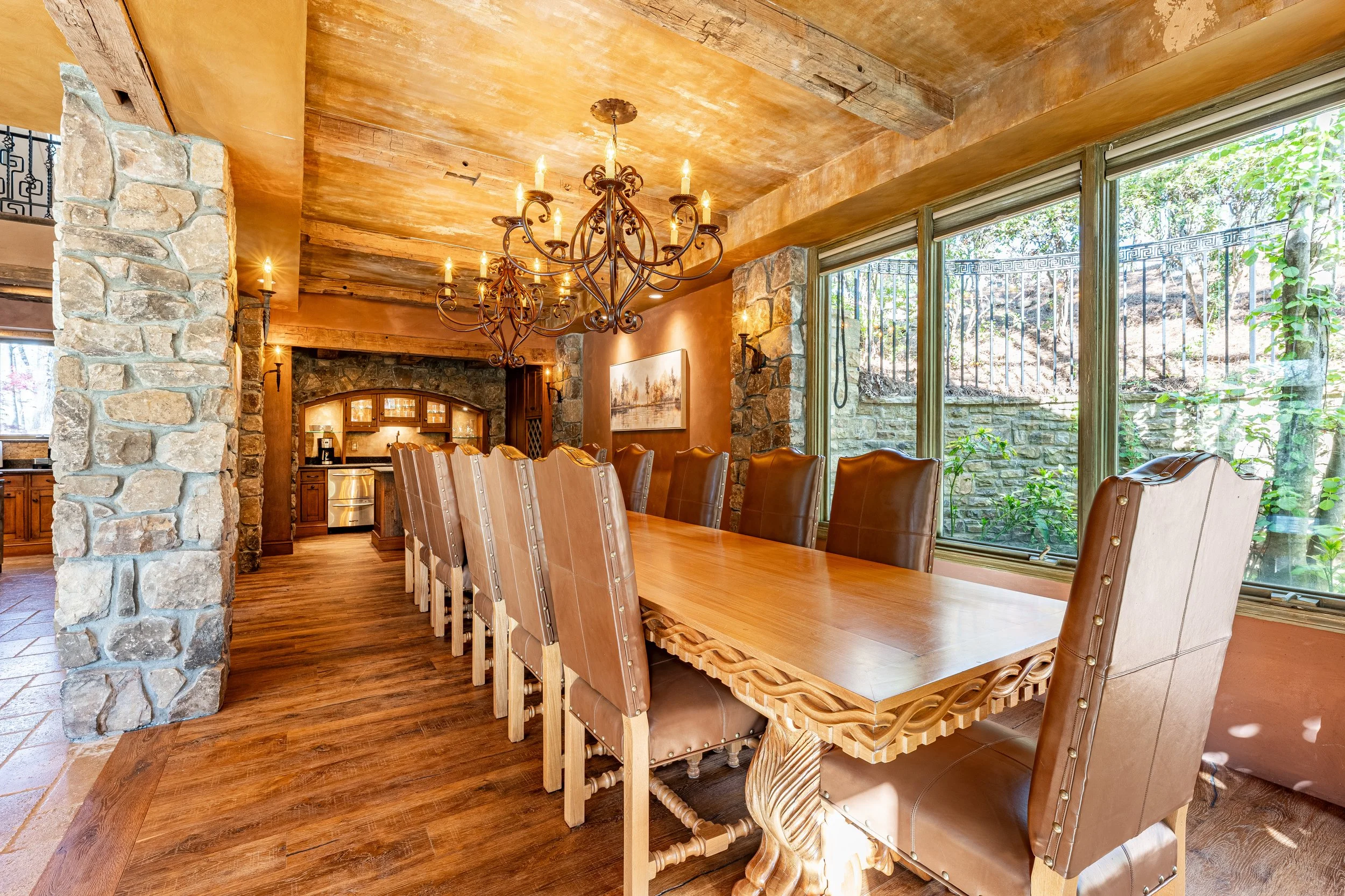 A spacious dining room with a long wooden table surrounded by eight high-back leather chairs, a wrought iron chandelier hanging overhead, large windows overlooking a garden, stone walls, and wooden accents.