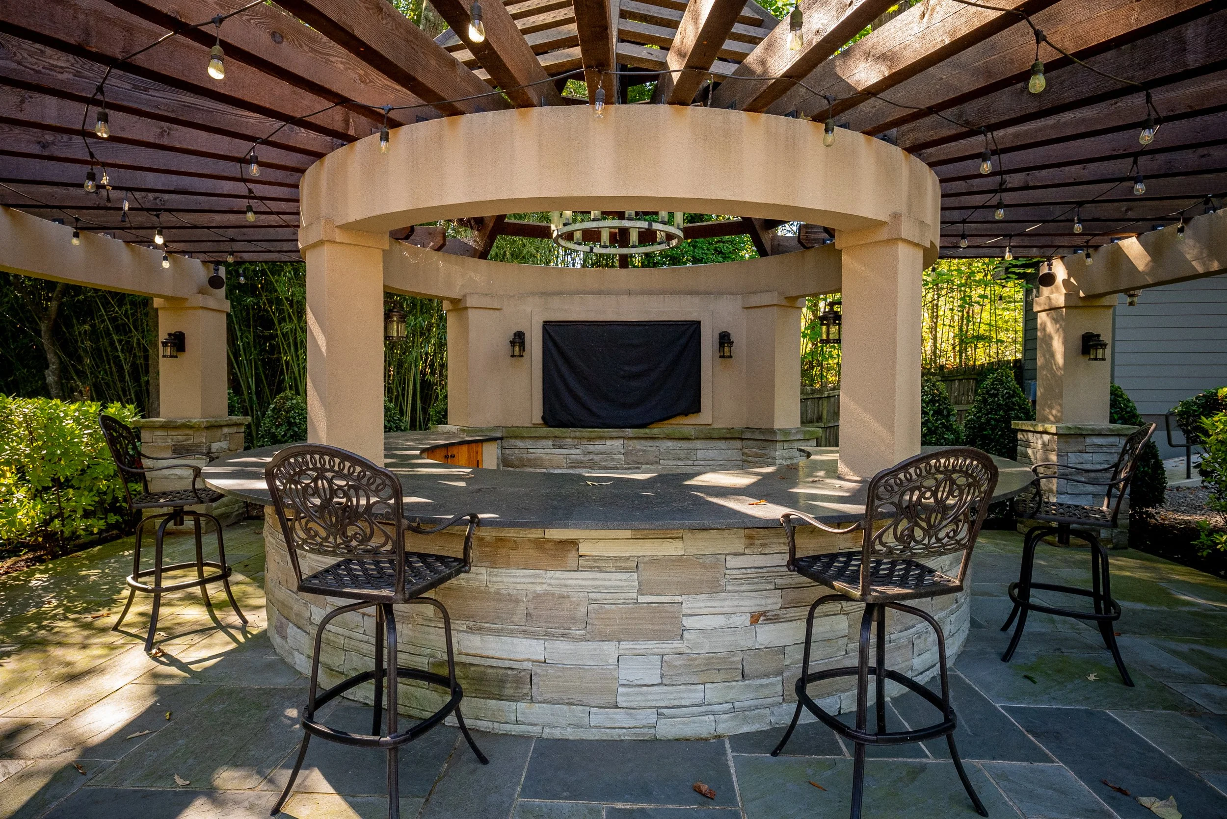 Outdoor patio with a circular bar area made of stone, surrounded by four metal bar stools, with a wooden pergola overhead decorated with string lights, and greenery in the background.