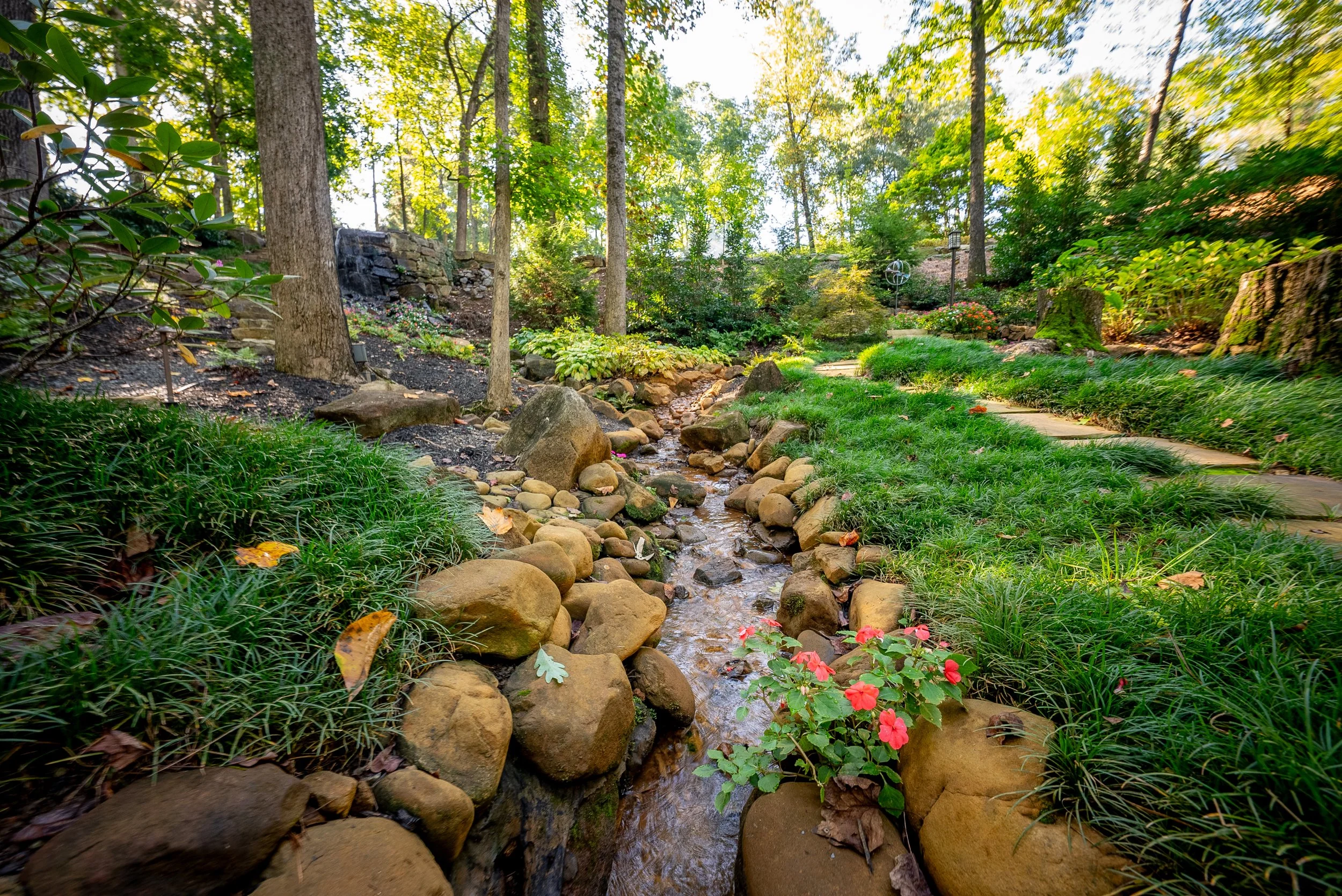 A serene garden with a small stream running through rocks, surrounded by lush greenery, trees, and flowering plants during daytime.