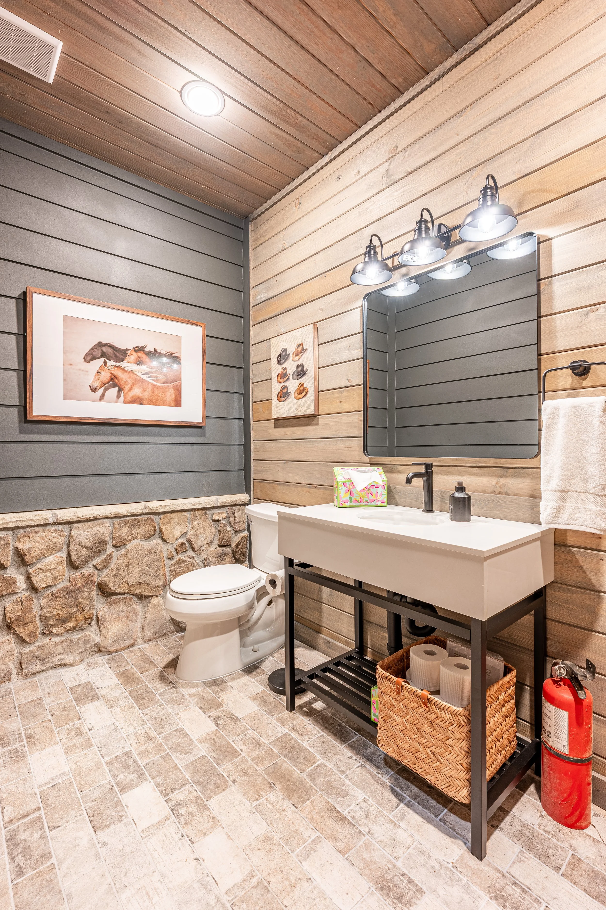 A rustic bathroom with wood-paneled walls, stone accents, and a beige tiled floor, featuring a white toilet, black metal vanity with a white basin sink, a large mirror, and decorative horse photos.