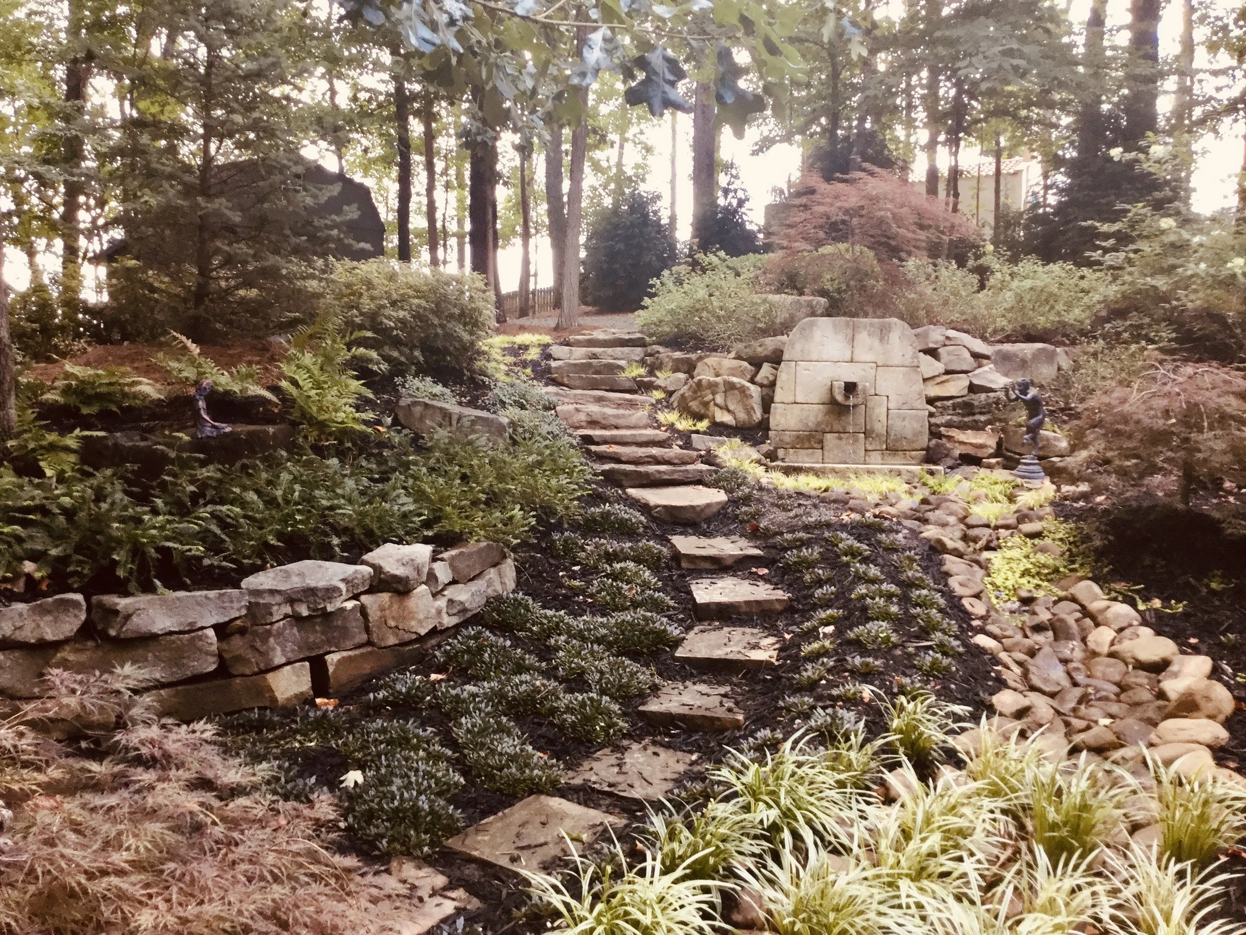 A landscaped garden with stone steps leading uphill, surrounded by various plants, shrubs, and trees, with a decorative stone fountain and small statues.