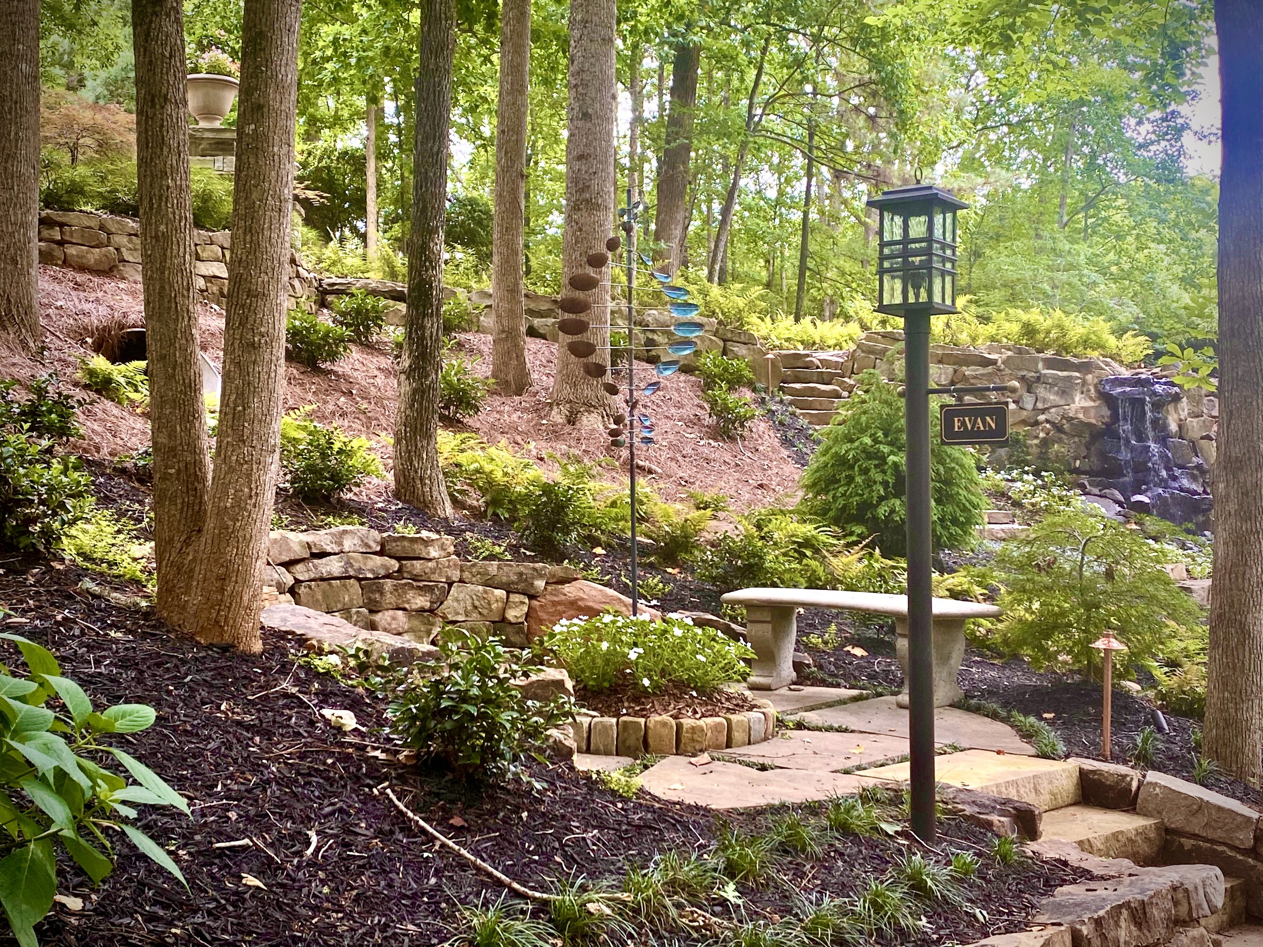 A landscaped garden with stairs, trees, ferns, and a water feature. There is a bench, a lamppost with a sign labeled 'Evan,' and decorative sculptures.