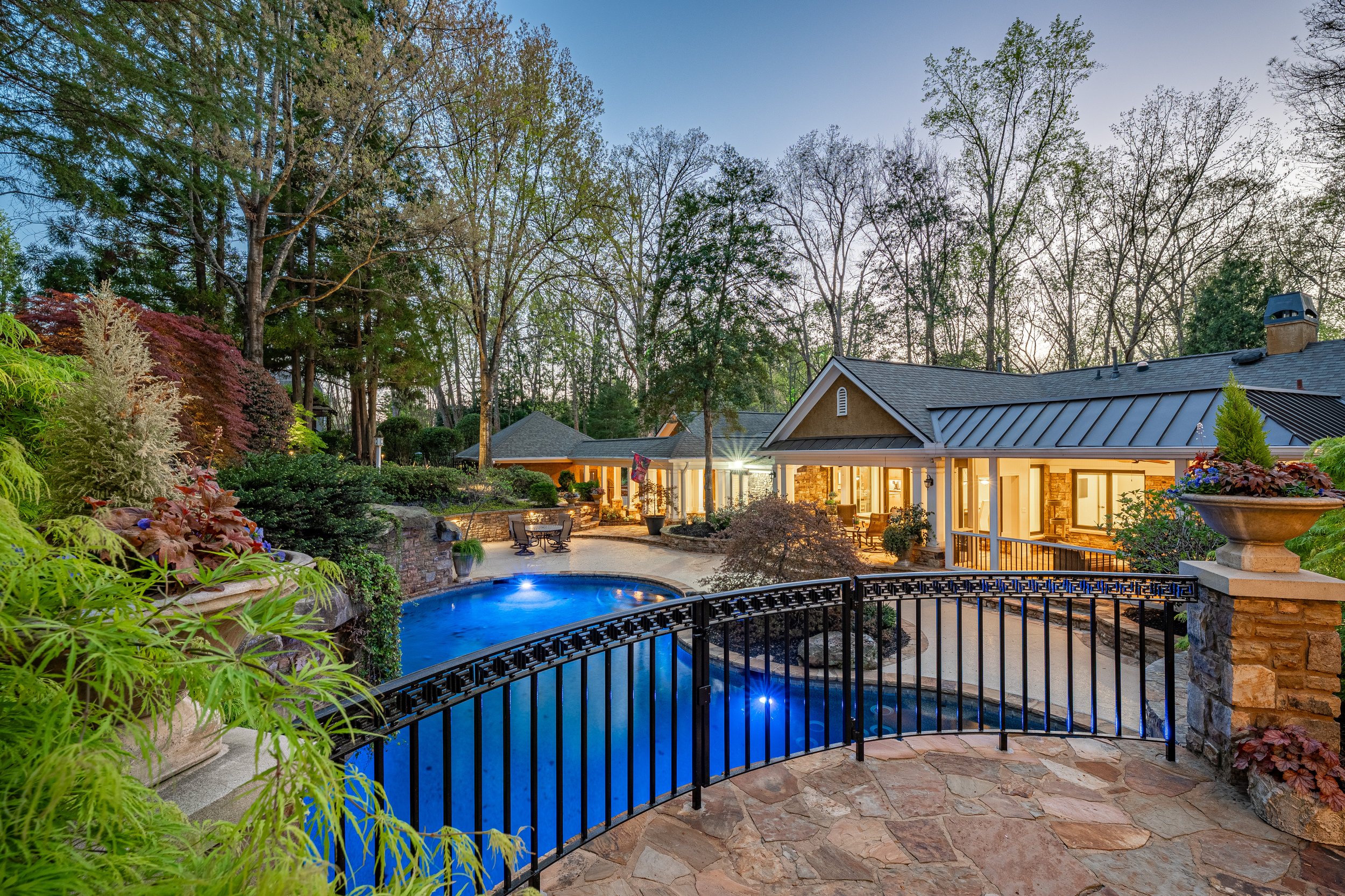 A backyard view featuring a swimming pool with blue lighting, surrounded by lush greenery, trees, and flowering plants. Adjacent to the pool is a house with large windows, warm lighting, and a covered patio area with outdoor furniture. The scene is captured during dusk with a clear sky and trees in the background.