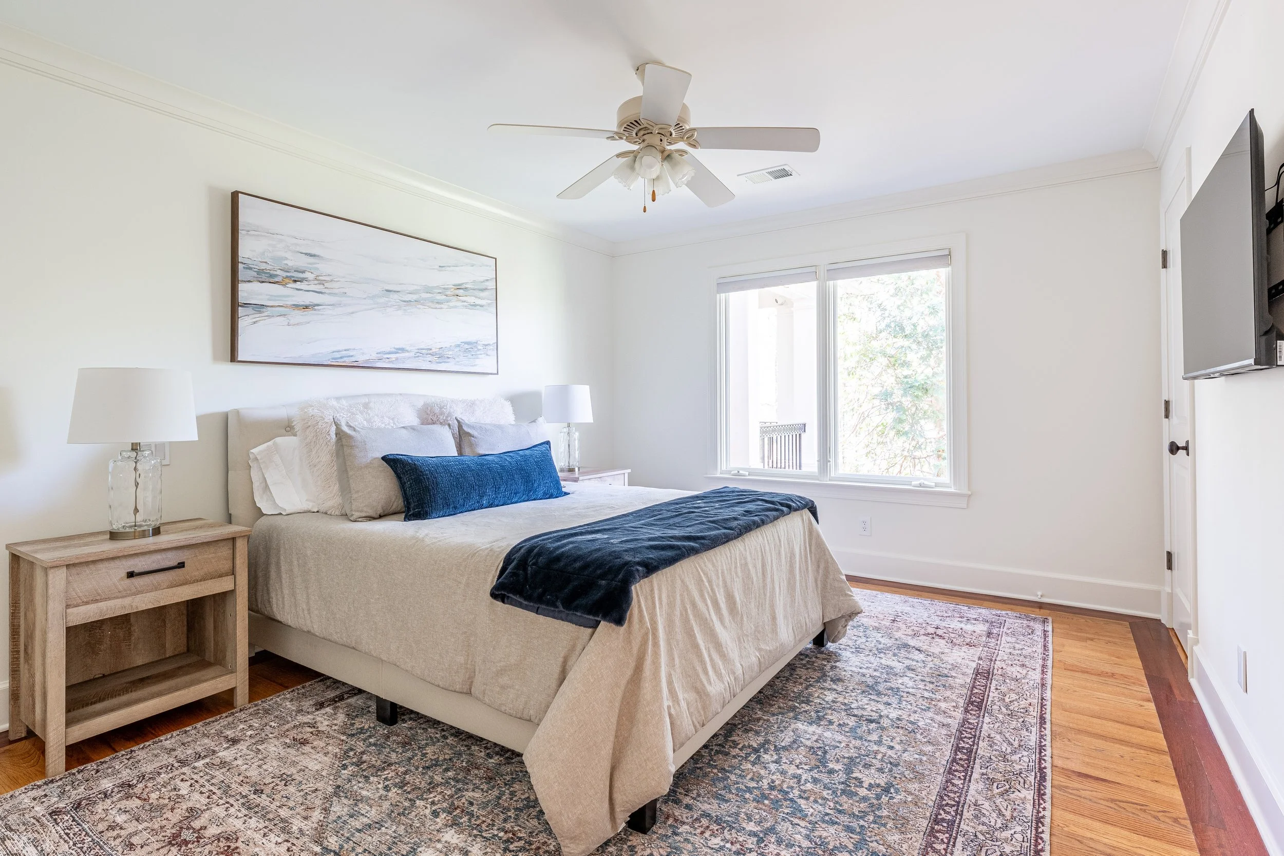 A bedroom with a bed, nightstands, table lamps, a ceiling fan, a window, and a mounted TV.
