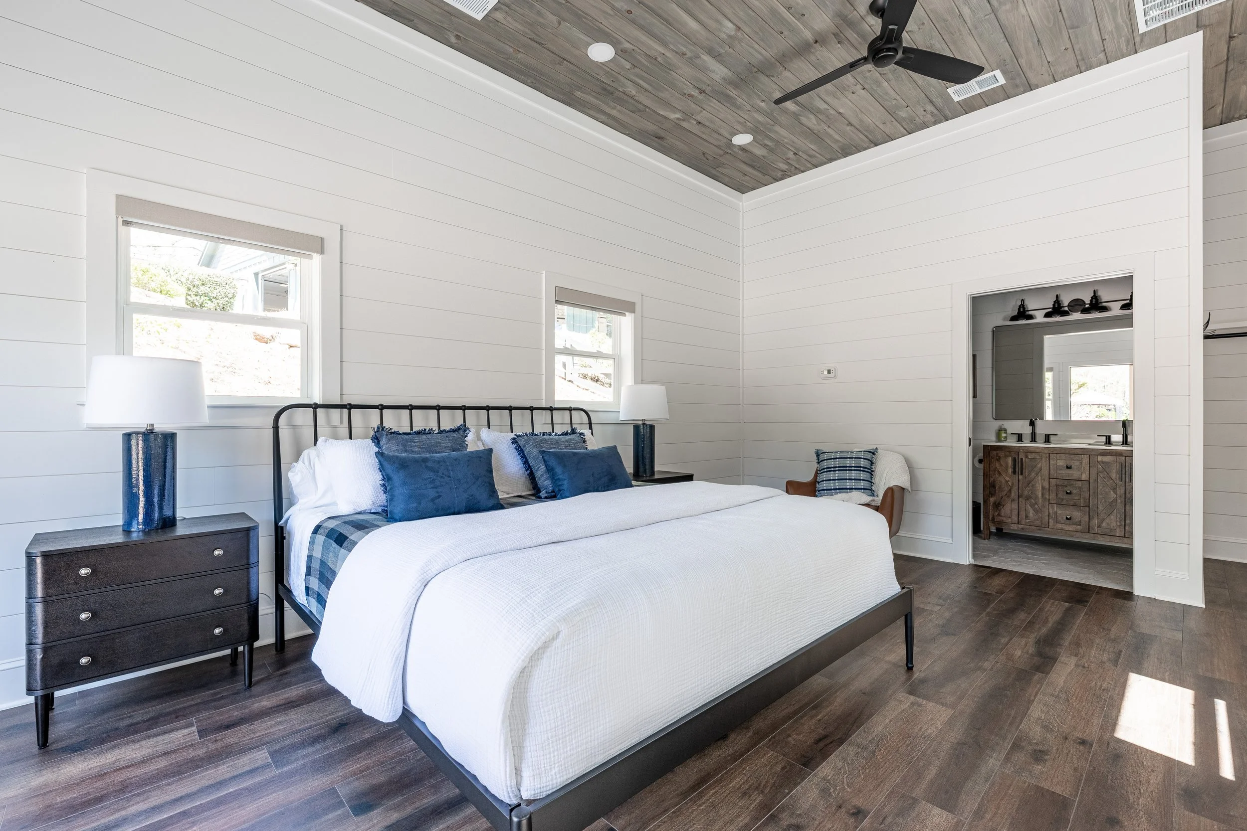 Bedroom with white shiplap walls, a black metal bed frame, white bedding, blue pillows, and wooden nightstands with lamps. Small windows and a view into an adjacent bathroom with a wooden vanity.