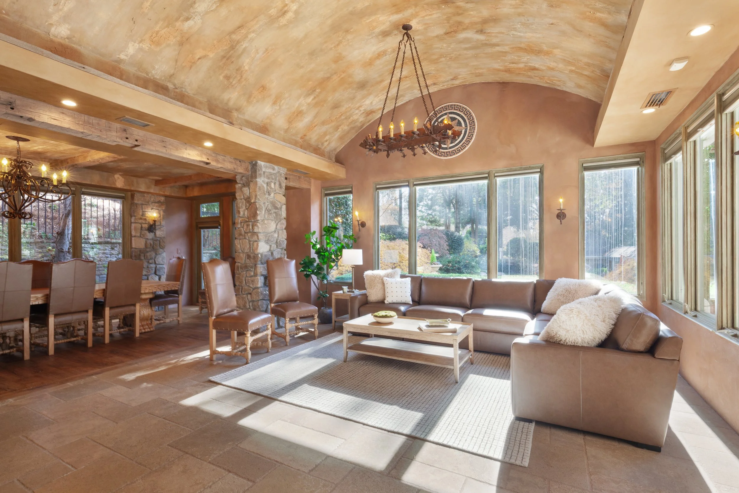 Well-lit living room with large windows, beige sectional sofa, fireplace, stone pillar, and rustic chandeliers.