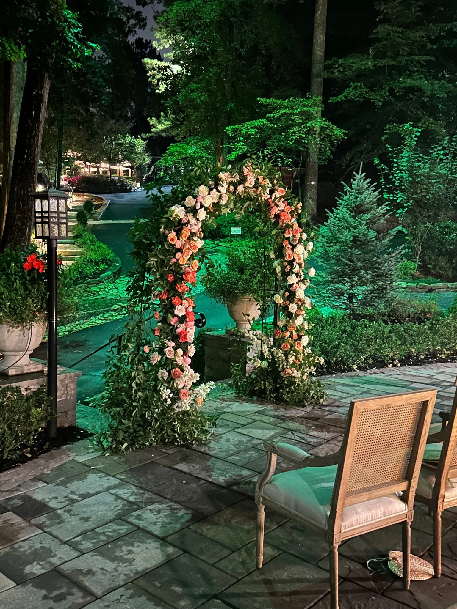 Nighttime outdoor garden scene with a floral arch decorated with pink and white roses, green trees, potted plants, a stone walkway, and a wooden chair on a patio.