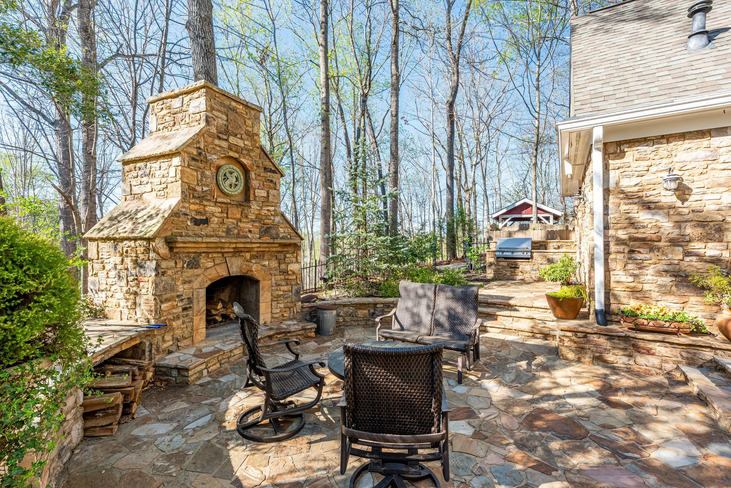 Outdoor stone patio area with a fireplace, seating, potted plants, and a barbecue grill, surrounded by trees.