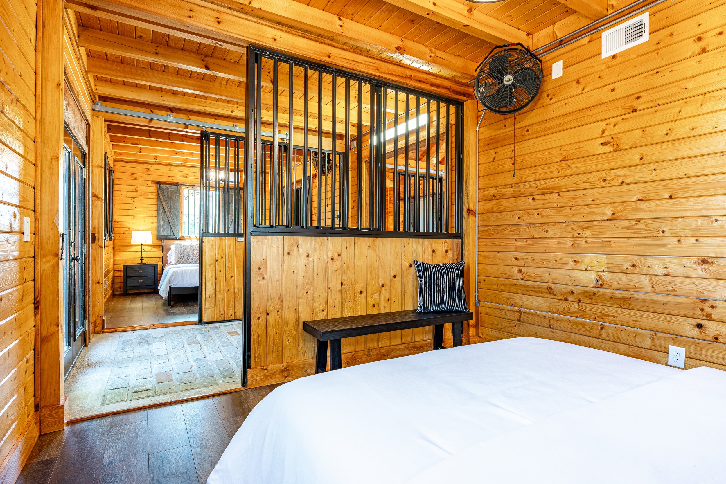 Interior of a cozy wooden cabin bedroom with a bed in the foreground, a bench with a striped pillow, a wall-mounted fan, and a semi-open partition showing a view into another bedroom with a nightstand and a lamp.