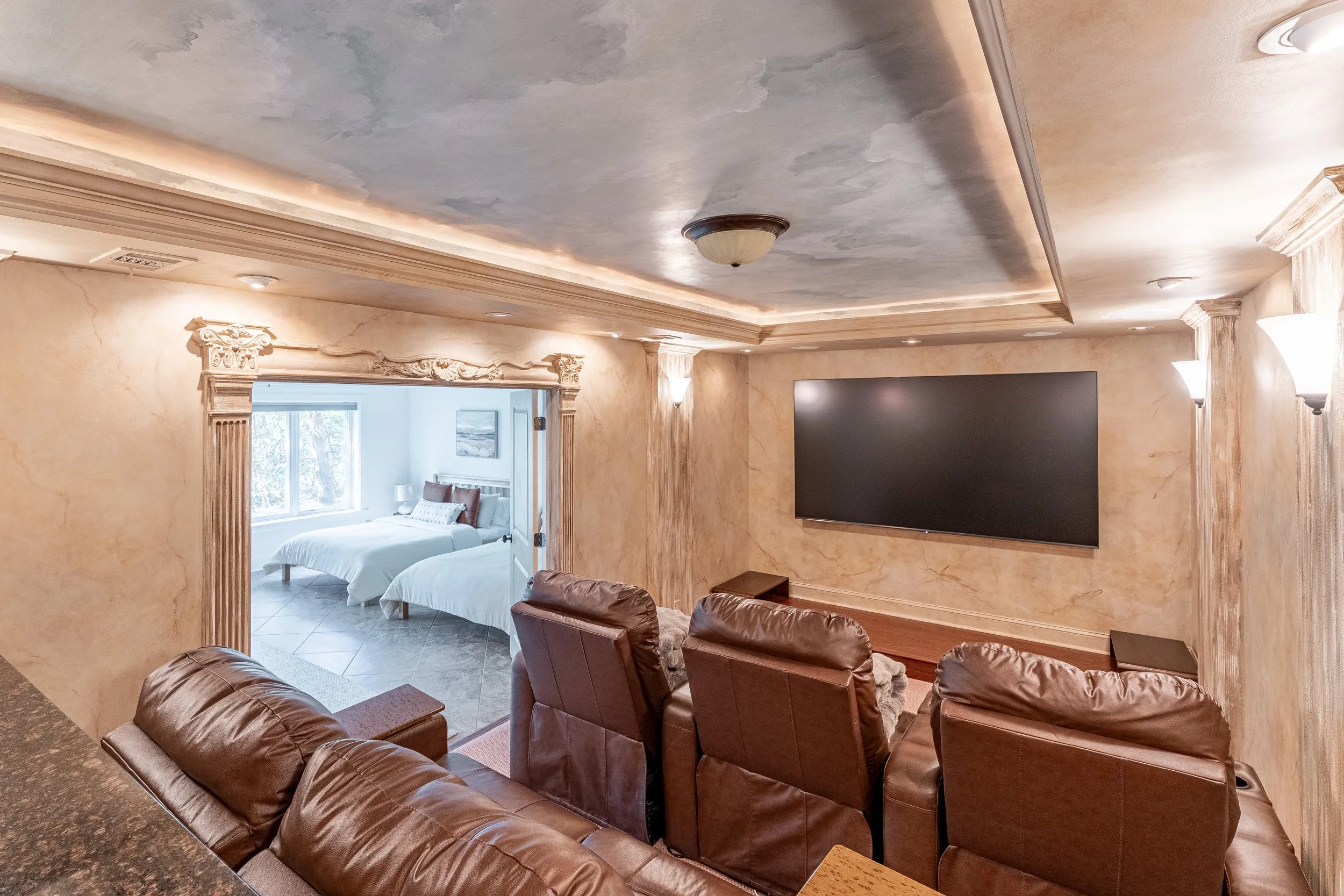 Living room with leather seating, a large wall-mounted TV, and a doorway leading to a bedroom with two beds, white bedding, and windows.