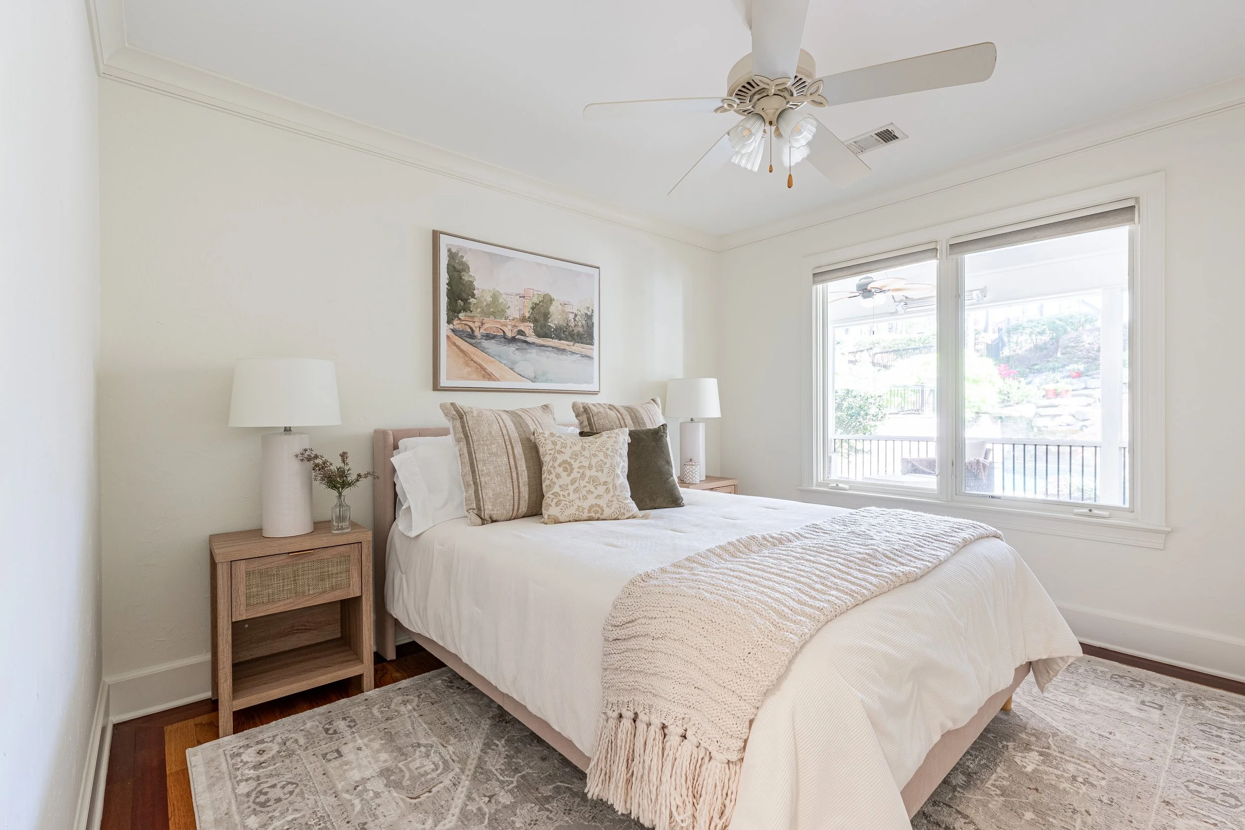 A neatly made bed with beige and patterned pillows in a bright bedroom with white walls, a window overlooking a porch, and a ceiling fan.