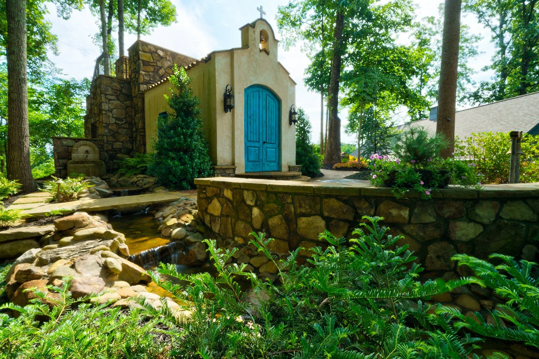 A small outdoor chapel with a blue door, surrounded by greenery and trees, with a stone wall and a garden fountain beside it.