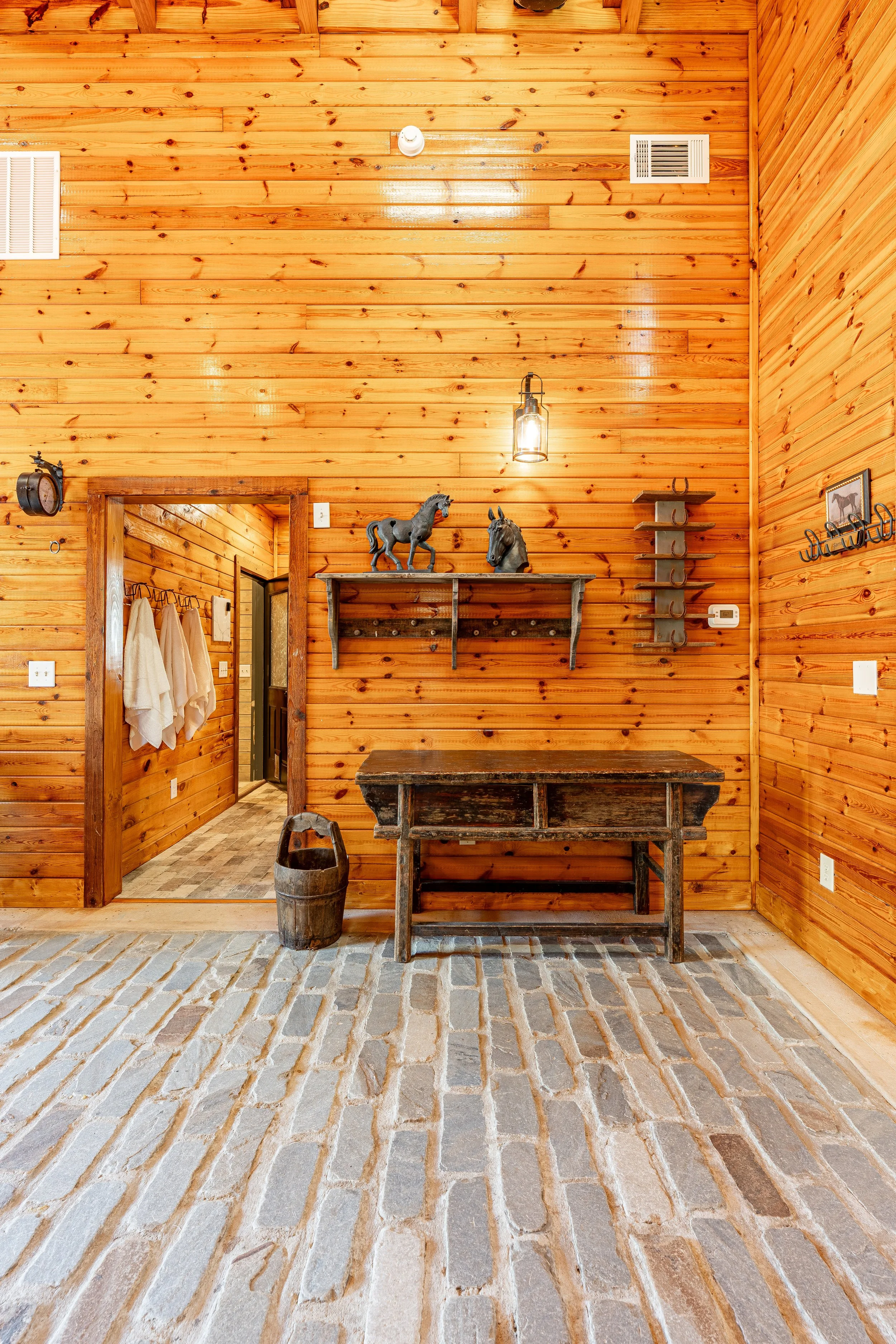 A rustic interior with wooden walls, a vintage wooden table, decorative horse figurines on a wall shelf, a hanging lantern light, and a doorway leading to a hallway with towels on hooks.