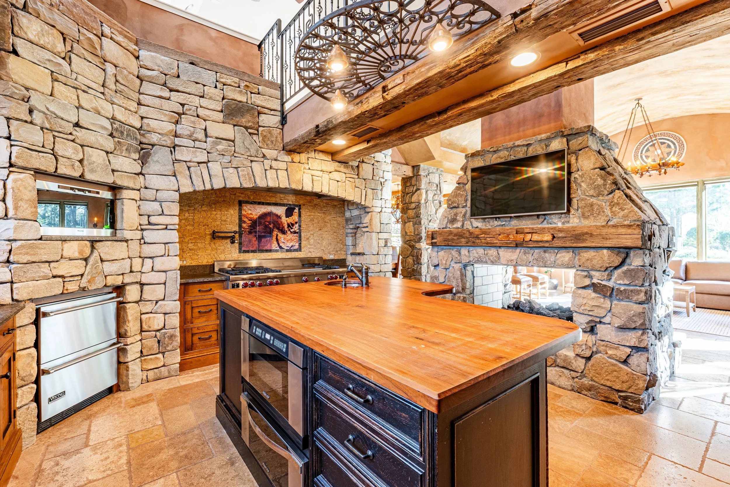 Rustic kitchen with stone walls, wooden cabinets, a large wooden island, and exposed wooden beams, featuring a stove, a wall-mounted TV, and a fireplace in the background.