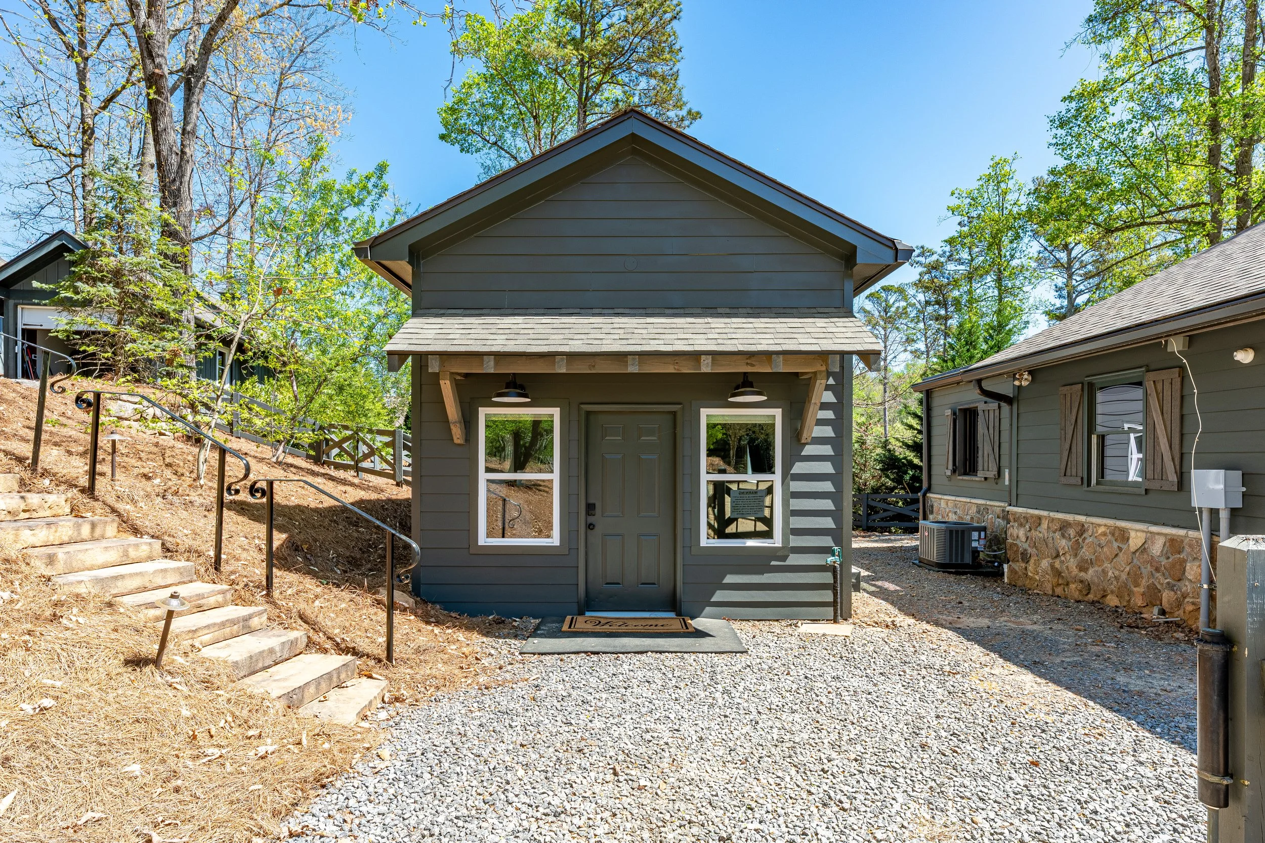 A small gray house with a gabled roof and front door, located between two other buildings, surrounded by trees, gravel driveway, and a set of stone steps leading up from the left.