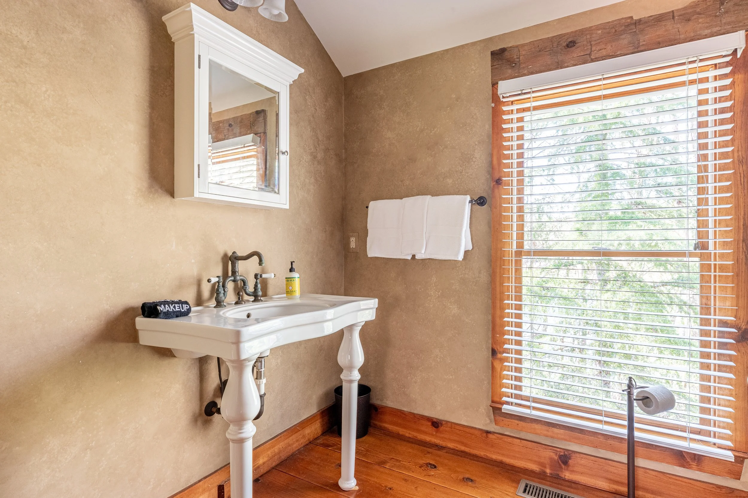 A rustic bathroom with a white porcelain sink, a small cabinet above it, a towel rack with white towels, a large window with blinds, and wooden floor and trim.