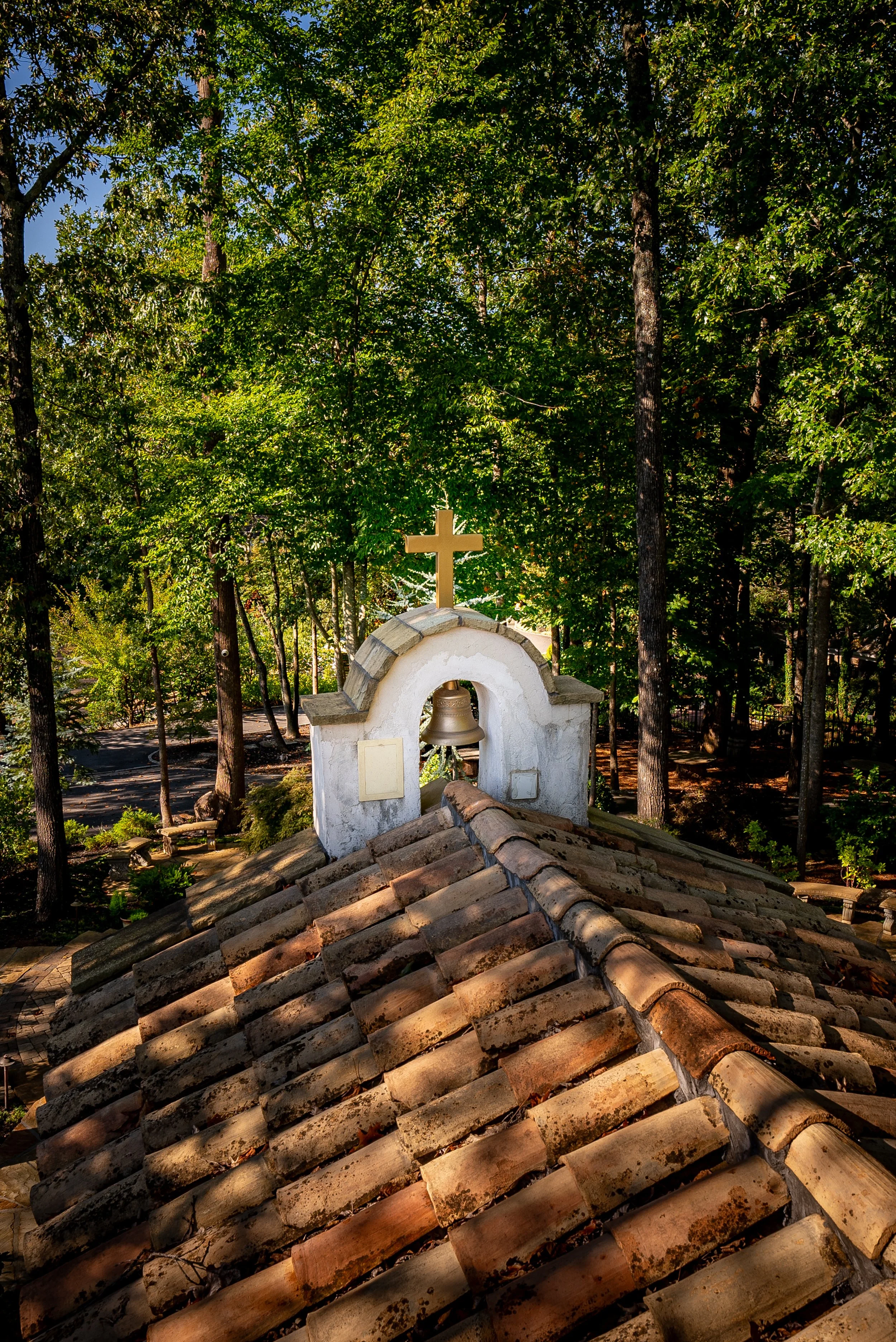 View of a church roof with a small bell tower topped with a cross, surrounded by tall green trees.