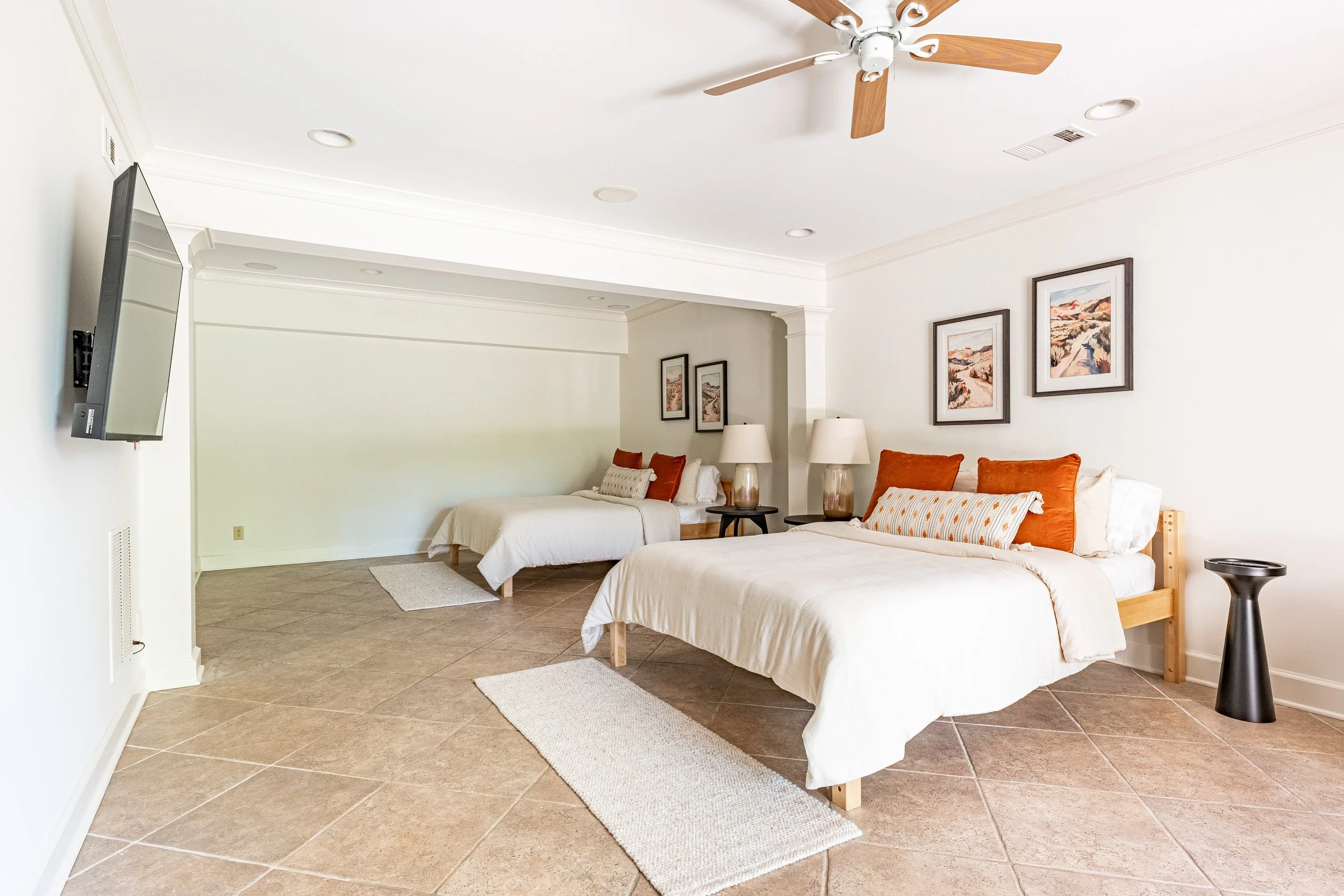 An interior view of a spacious bedroom with two beds, one larger and one smaller, with white bedding and orange and patterned pillows. There are three framed landscape pictures on the wall above the larger bed. Two lamps are on small tables between t