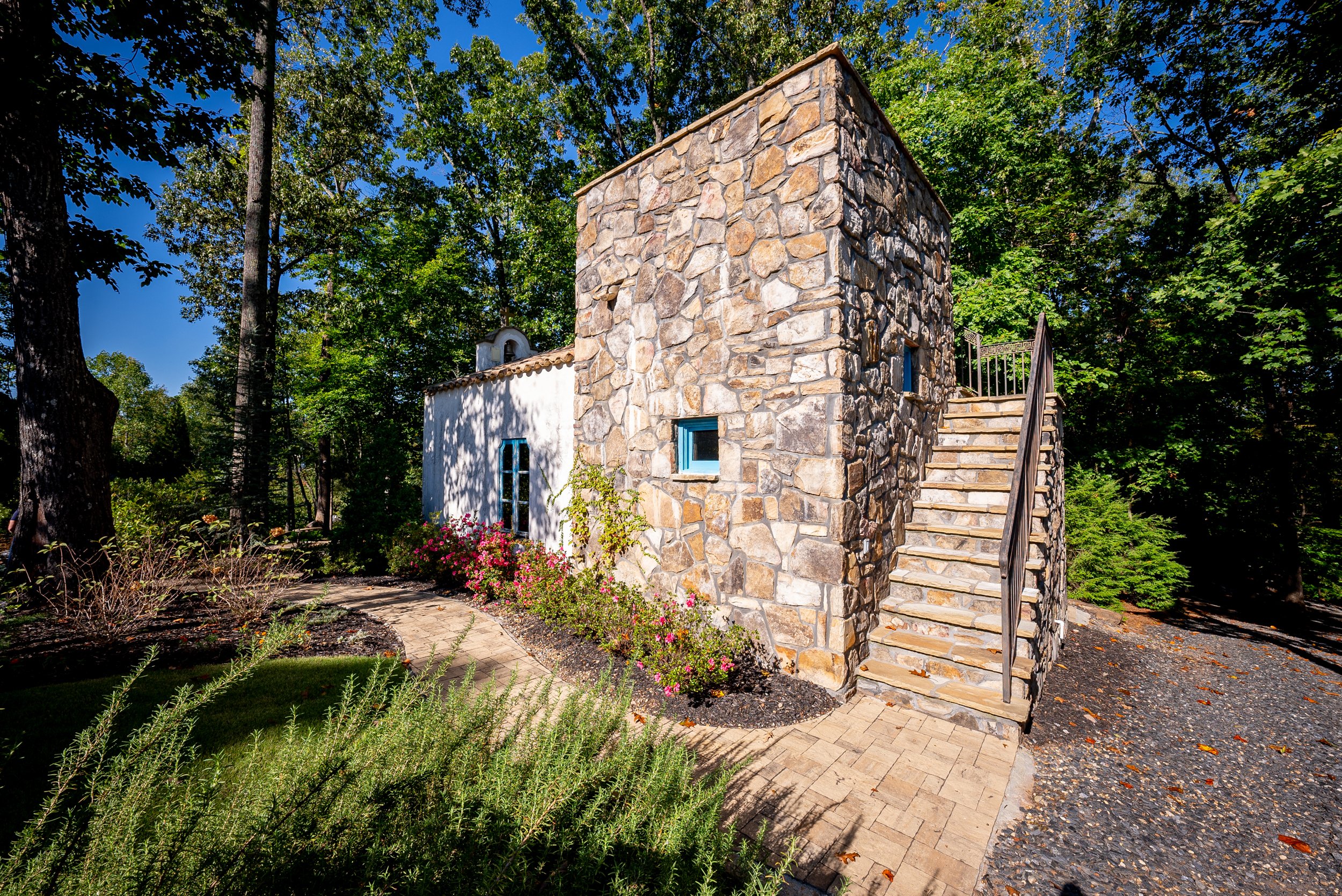 A house with a stone exterior on one side and white walls on the other, featuring a staircase with a metal railing and small windows, surrounded by greenery and a paved walkway.