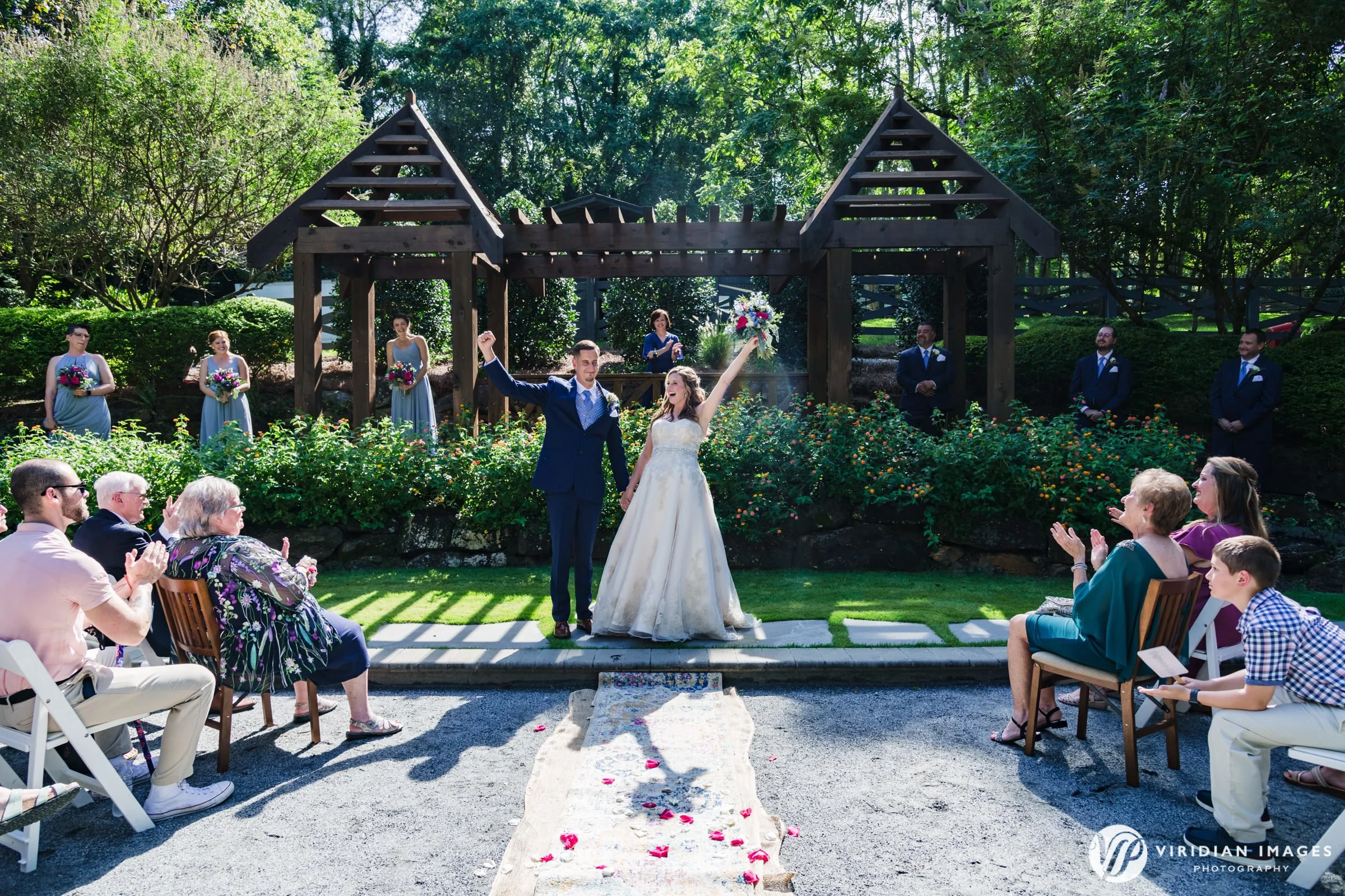 A wedding ceremony outdoors with a bride and groom holding hands in the center, celebrating under a wooden arch, surrounded by guests and bridesmaids and groomsmen, on a sunny day with lush greenery in the background.