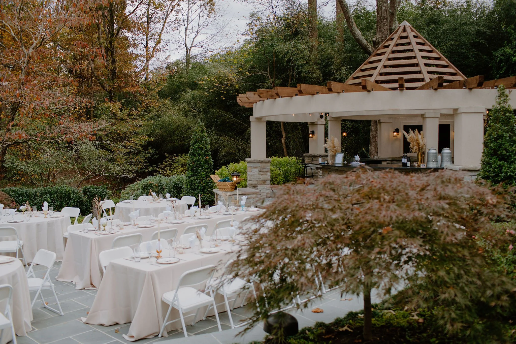 Outdoor event setup with white tables and chairs, decorated with white tablecloths, in front of a pavilion with a peaked roof, surrounded by trees with autumn leaves.