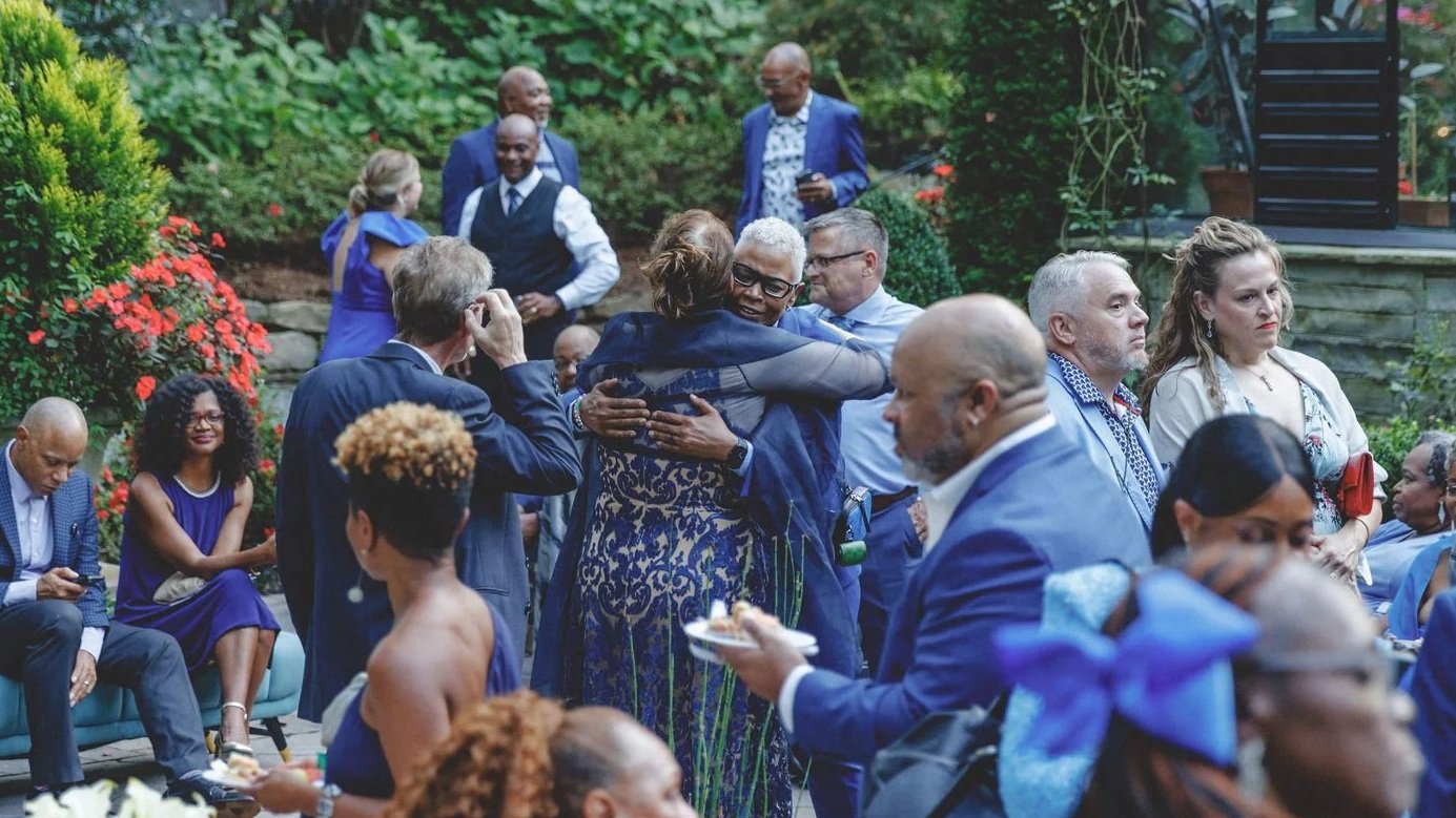 People at a social gathering hugging and conversing outdoors in a garden setting with colorful flowers and greenery.