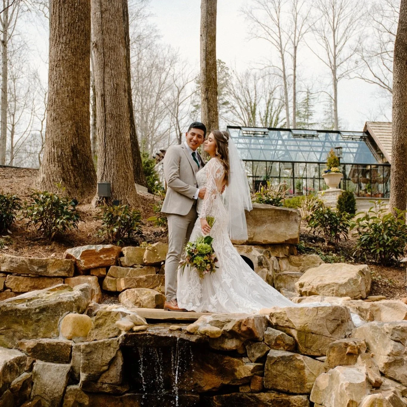 A bride and groom in wedding attire standing on rocks near a small waterfall, smiling and embracing outdoors with trees and a greenhouse in the background.